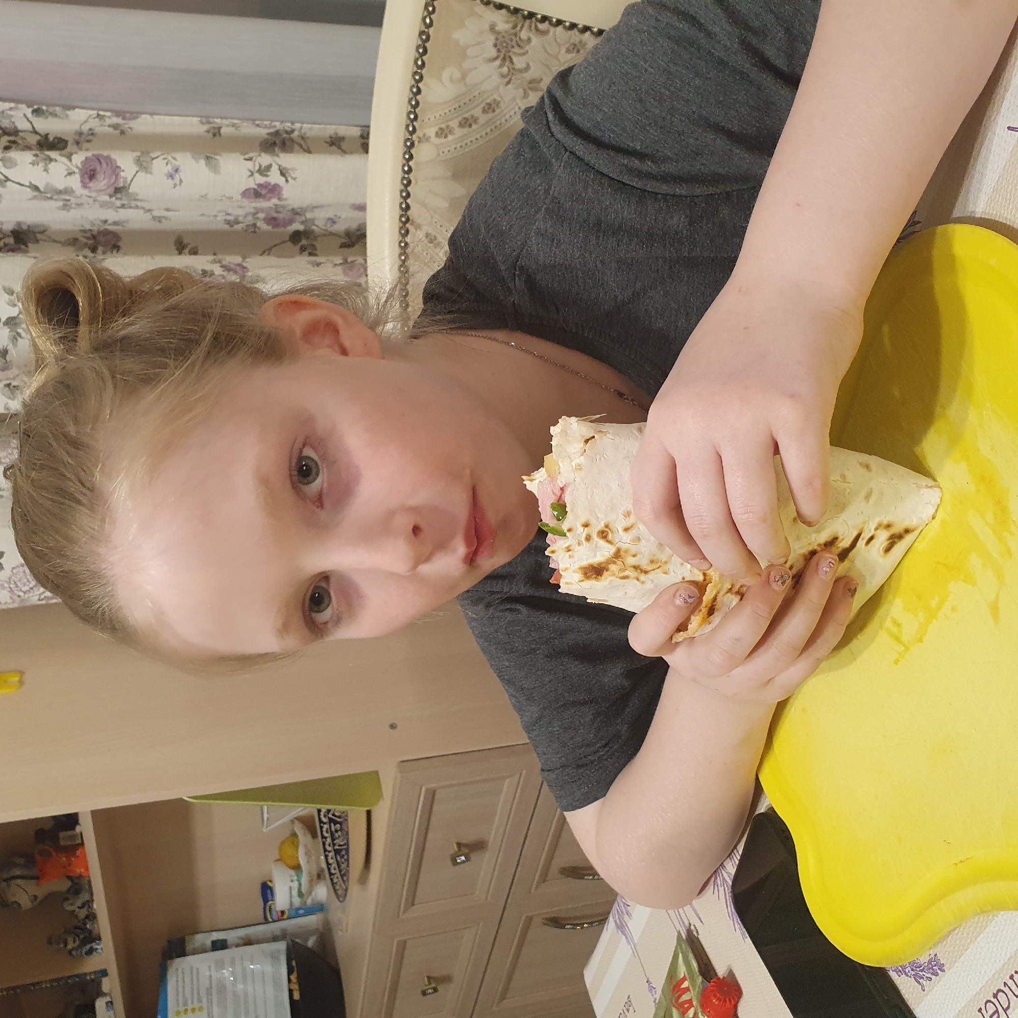 An indoor, eye-level shot captures a young individual, likely a female, holding and eating a large wrap or shawarma. The person, with fair skin, light blonde hair tied up, and light-colored eyes, is positioned at a table, looking directly at the viewer with a neutral expression, mid-bite from the wrap. They are wearing a dark grey short-sleeved t-shirt and a delicate chain necklace. Their fingernails are painted with small, light-colored designs, possibly butterflies or hearts.
The wrap itself appears to be made from a lightly grilled tortilla, filled with ingredients such as pinkish ham or sausage, green leafy vegetables, and other light-colored components like cheese or sauce. A bright yellow, irregularly shaped cutting board is placed on the table next to the wrap.
The scene is set in what appears to be a home dining area or kitchen. To the left of the person are light-colored wooden cabinets or shelves, displaying various packaged items and a yellow lemon. To the right, a cream-colored chair with decorative metallic studs and an embroidered floral pattern is visible. The table is covered with a white and purple patterned tablecloth, featuring what looks like lavender motifs. Partially visible text on the tablecloth reads "LAVENDER". A small red, strawberry-shaped object, possibly a decoration or dish, is also on the table, with the letters "MA" visible on it. In the background, floral curtains with purple rose patterns can be seen. The lighting suggests an indoor setting without strong natural light sources. The location context is Chaikovsky, Russia.