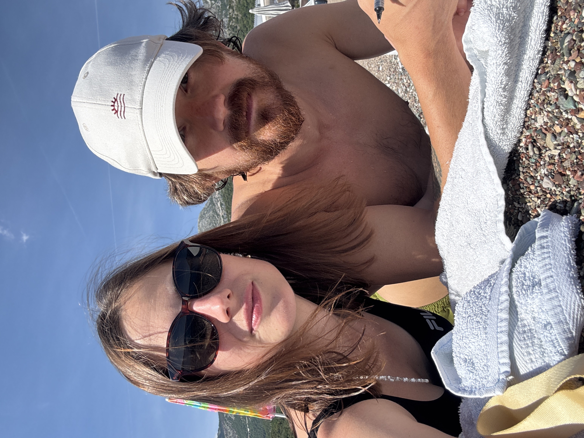 Two individuals, a woman and a man, are captured in a selfie taken outdoors on a sunny day, likely at a beach in Boreti, Montenegro. The woman, on the left, has medium-length brown hair and is wearing dark sunglasses and a black swimsuit. A beaded necklace hangs over her swimsuit. The man, on the right, wears a white baseball cap and has a beard and mustache. He is shirtless. Both are lying down, with the woman's head resting on her arm and the man's arm visible in the foreground. White towels are spread out on the pebble-strewn ground, and part of a colorful umbrella can be seen behind the woman. In the background, mountains and the sky are visible.