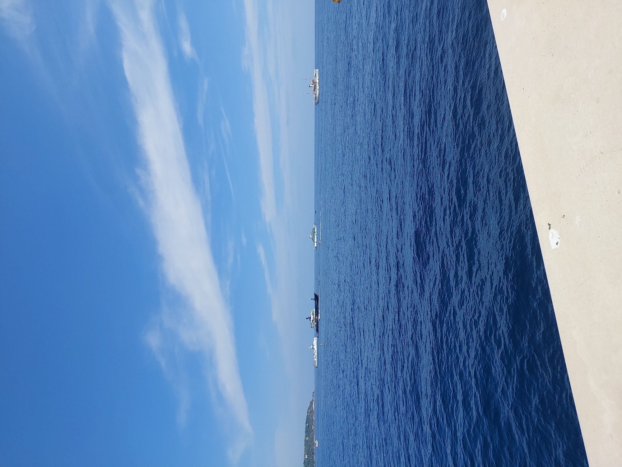 The image depicts a wide view of the Mediterranean Sea off the coast of Monaco. The water is a deep, vibrant blue with small waves. In the distance, several large yachts and a few smaller boats are visible on the horizon. To the far left, a lush green coastline with buildings can be seen, suggesting the proximity of land, likely Monaco's famed coastline. The sky is a clear, bright blue with thin, wispy white clouds scattered across it. In the foreground, a light-colored concrete structure, possibly a quay or promenade, runs along the bottom of the frame, hinting at the viewpoint being from the edge of the city's harbor or waterfront. The scene conveys a sense of calm and luxury, characteristic of Monaco.