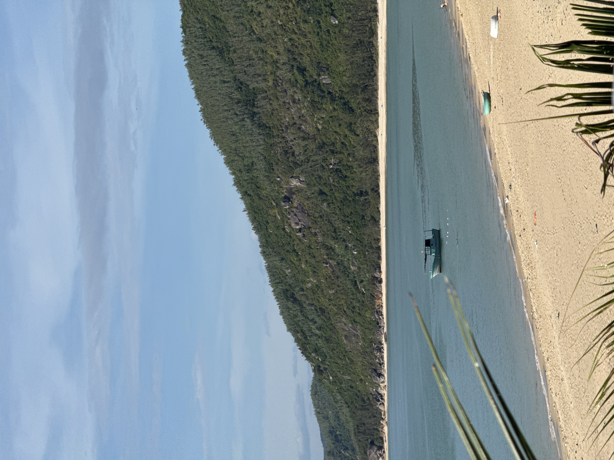 The image depicts a tranquil beach scene in Song Cau, Vietnam. A calm, turquoise body of water stretches towards a lush, green, tree-covered mountainside that forms the backdrop. In the foreground, a wide, sandy beach is visible with a few small boats and what appear to be beach umbrellas or shelters scattered across it. A single, green and blue fishing boat is anchored or drifting in the shallow water near the shore. The sky above is a clear blue with a few wispy clouds. The overall impression is one of a serene, natural coastal environment.