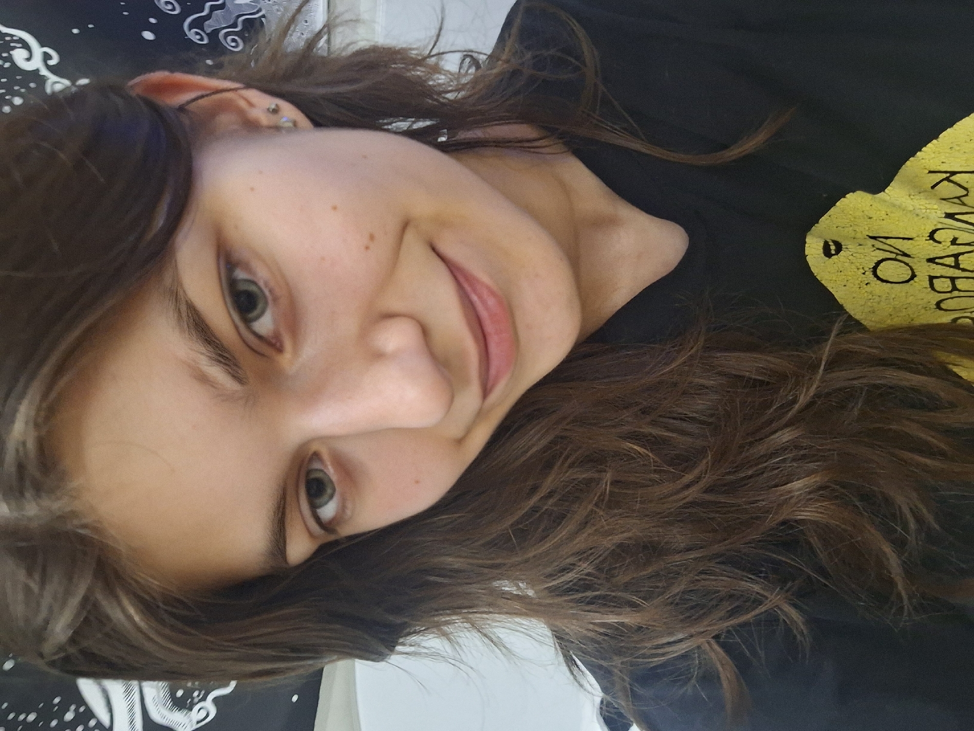 The media file is a close-up portrait of a young woman, captured in what appears to be a casual indoor setting. Her head is significantly tilted to the left, giving the image a dynamic, candid feel, possibly indicating a selfie. She has long, wavy dark brown hair that frames her face and partially covers her shoulders.
The woman is looking directly at the camera with light-colored (likely green or hazel) eyes and a subtle, closed-mouth smile, conveying a relaxed and pleasant demeanor. Her complexion is fair, with a few visible freckles on her cheek. A silver ear piercing is visible on her left ear cartilage. She is wearing a black t-shirt.
In the background, behind her head, there's a black surface with abstract white swirl patterns and dots, possibly a pillow, blanket, or tapestry. To her right, on her black t-shirt, there is yellow text written in Cyrillic script. The visible words, when oriented correctly, read "ОЙ КВА КУМАК". The phrase "ОЙ КВА" (Oy Kva) is a colloquial Bulgarian expression meaning "Oh, what a...". The word "КУМАК" (Kumak) is not a standard Bulgarian word and might be a name, slang, or a specific reference. The overall impression is a personal and informal photograph.