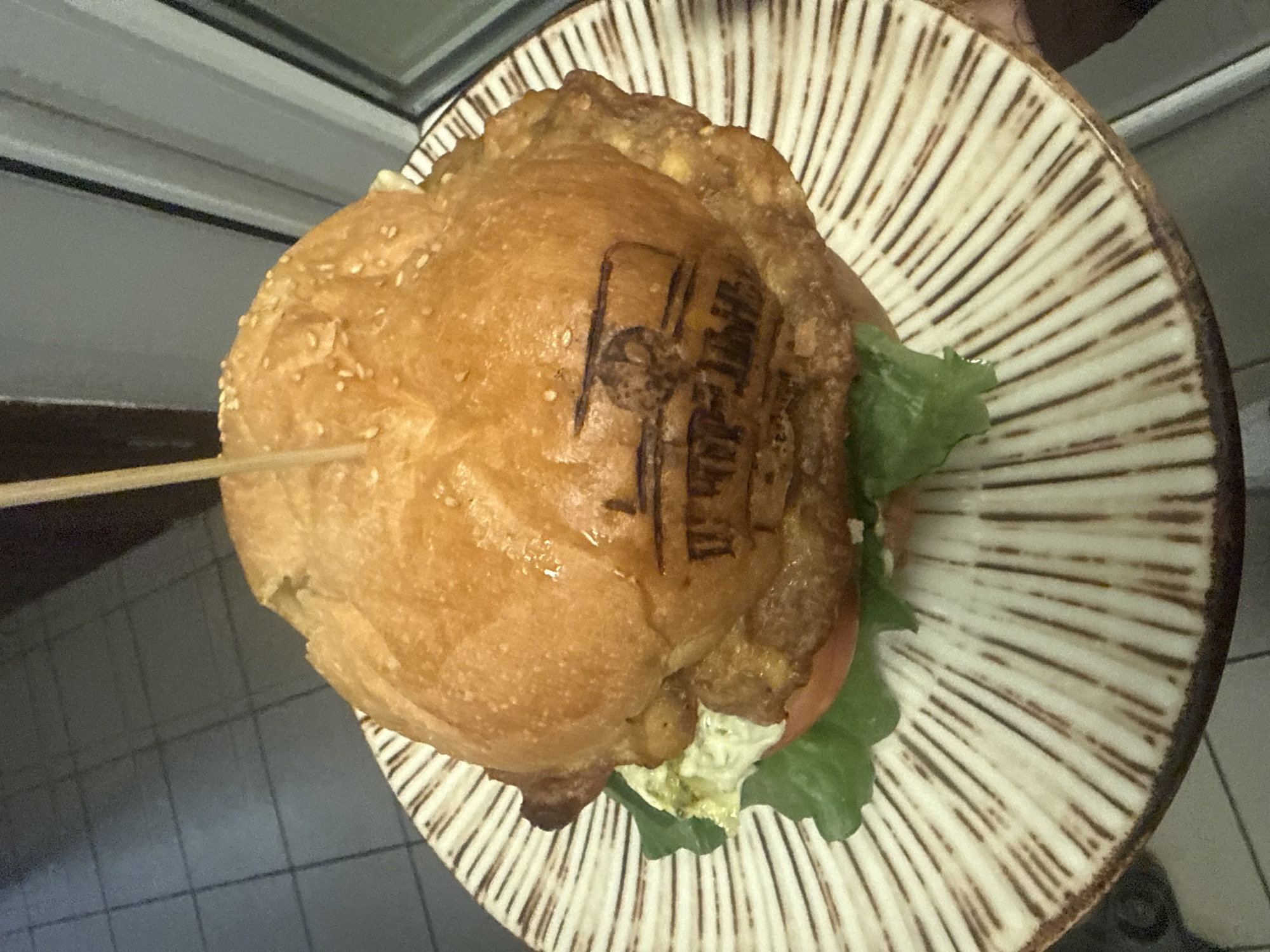 This media file presents a top-down view of a large burger served on a distinctive plate, set indoors in Birobidzhan, Russia. The central subject is the burger, characterized by a golden-brown, sesame-seed-topped bun. Beneath the top bun, a substantial fried layer is visible, possibly a fried egg or a battered, crispy patty. Green lettuce and a slice of red tomato are peeking out from the sides, contributing to the burger's height. A wooden skewer pierces through the center, helping to hold the components in place.
A notable detail is the dark, branded logo or text imprinted on the top bun. While challenging to fully decipher from the image, it appears to be a stylized, multi-lined graphic, likely representing the establishment's name or a signature item. The burger rests on a round, light-colored ceramic plate with prominent radial brown stripes, creating a sunburst-like pattern.
The background reveals a light-colored, tiled floor with dark grout lines, along with a dark edge that suggests a counter or table. The scene depicts the presentation of food, implying it is ready to be consumed or has just been served. No people are visible, and the lighting suggests an indoor setting.