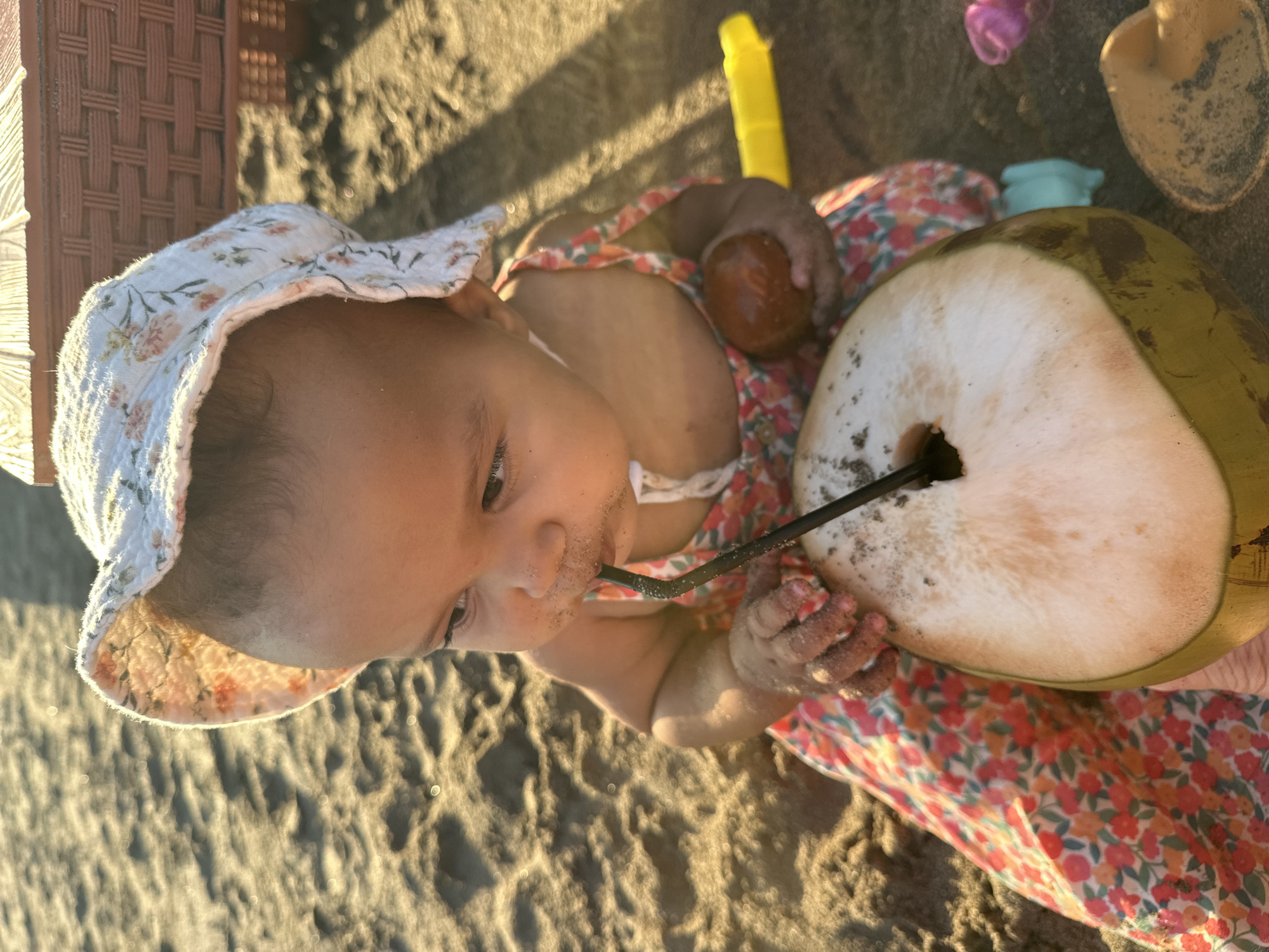 In North Kuta, Indonesia, a close-up shot captures a young child sitting on sandy ground, wearing a floral print sun hat and a matching dress. The child is holding a halved coconut with a straw inserted, appearing to drink from it. The child's hands are covered in sand, and a small, brown, round object is held in the child's right hand. In the background, a reddish-brown woven object and a yellow plastic toy are partially visible. The sandy environment suggests a beach or similar outdoor setting.