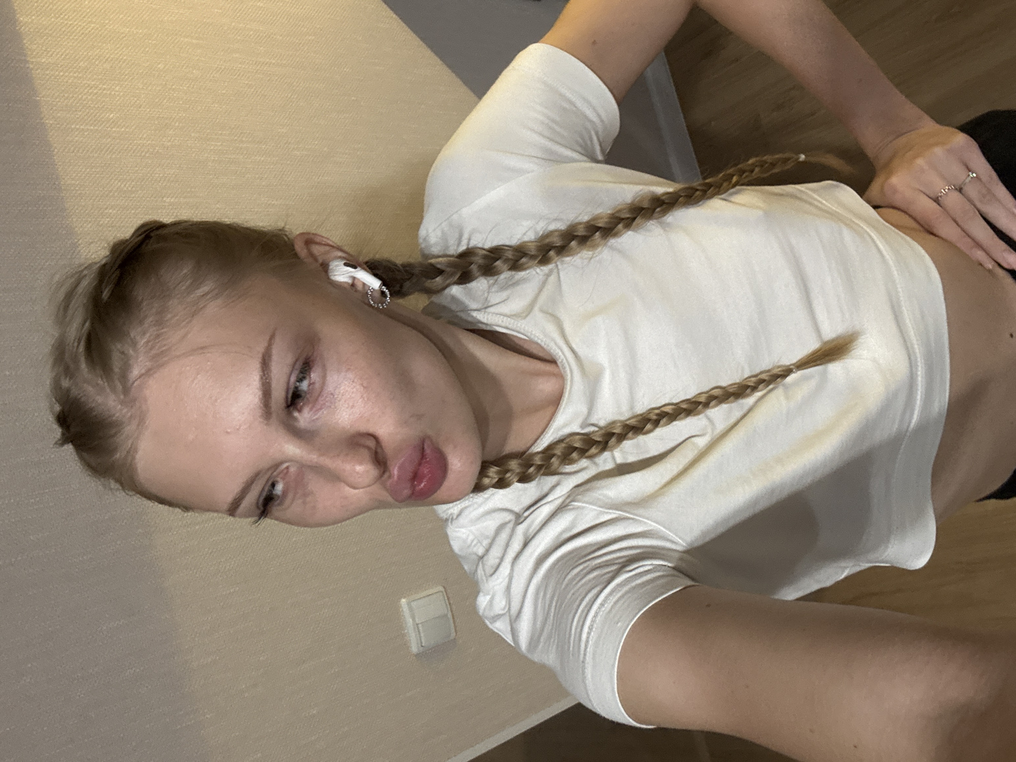 A young Caucasian woman with blonde hair styled in two braids is posing for a selfie in a dimly lit room. She is wearing a white cropped t-shirt and has white wireless earbuds in her ears. Her lips are pursed in a duckface pose. The background consists of textured walls and a wooden floor. A light switch is visible on the wall. The scene does not provide any specific geographical markers to indicate a location in Perm, Russia.