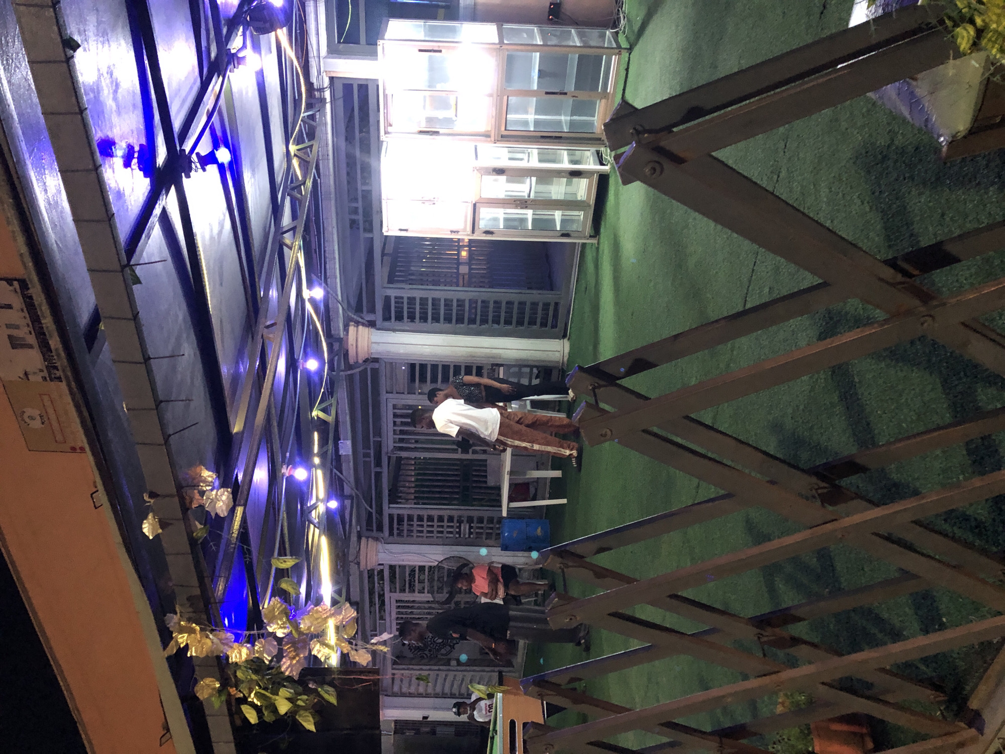 A low-angle, rotated outdoor shot taken at night depicts a gathering of people in Ado, Nigeria. The scene is lit by blue string lights attached to a metal pergola-like structure. A man wearing a white t-shirt and brown pants stands near a table. A small group is gathered in the background. Stacks of window frame-like structures rest on the right. Artificial turf covers the ground. A portion of a building is visible on the left. Metal scissor-like construction elements are featured prominently.