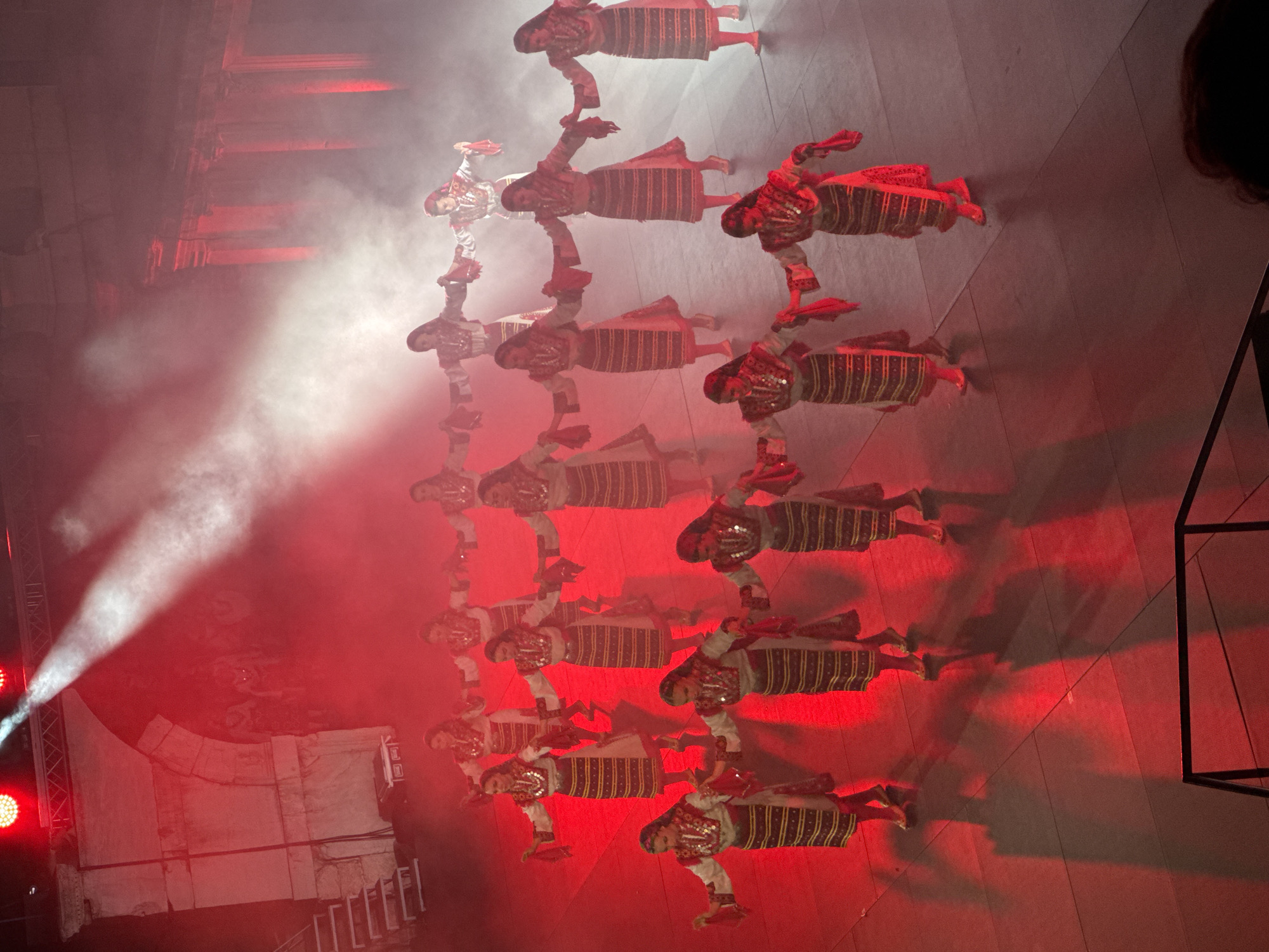 A group of approximately fifteen women in traditional folk costumes are performing a dance on a stage under red lighting and smoke effects. The women are arranged in multiple rows and appear to be holding hands and dancing in a circular or semi-circular formation. Their costumes consist of white blouses with embroidered yokes, striped skirts, and headscarves. The stage is dark with red floor lighting casting long shadows. In the background, there is a large stone archway and pillars, suggesting an ancient or historical architectural setting. The lighting is dramatic, with spotlights creating beams through the smoke.