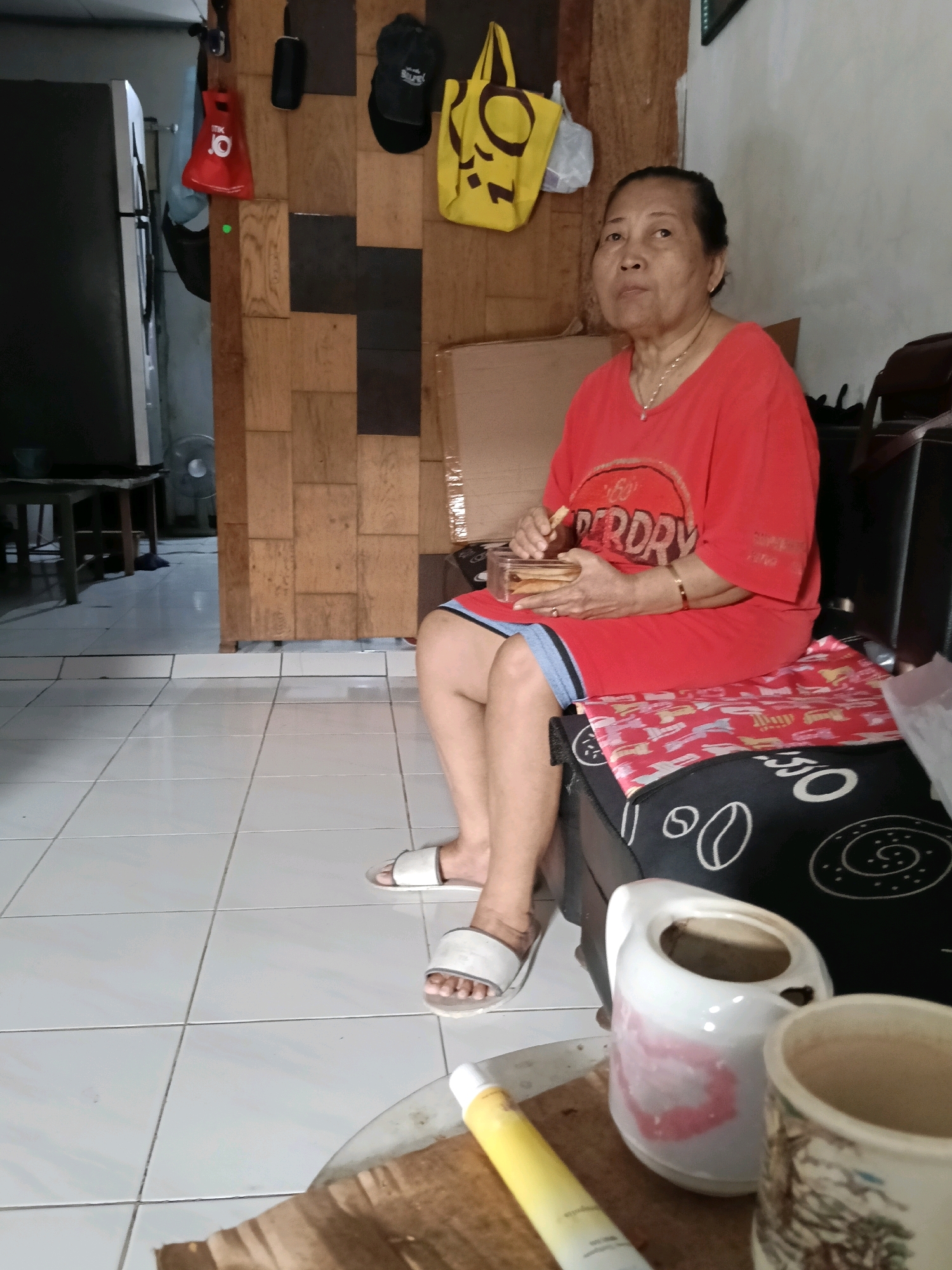 An elderly Asian woman is seated on a dark cushion, wearing a red t-shirt and shorts, holding a clear plastic container of crackers. She is looking to her left, with a fork in her right hand. The woman is wearing white slides on her feet and a gold bracelet on her left wrist. In front of her, on a table, are a white pitcher with pink markings and a mug with a decorative design. The background shows a wall paneled with wood, a yellow tote bag hanging, and a black baseball cap. The scene appears to be indoors, possibly a home in Jatimulya, Indonesia, with white tiled flooring.