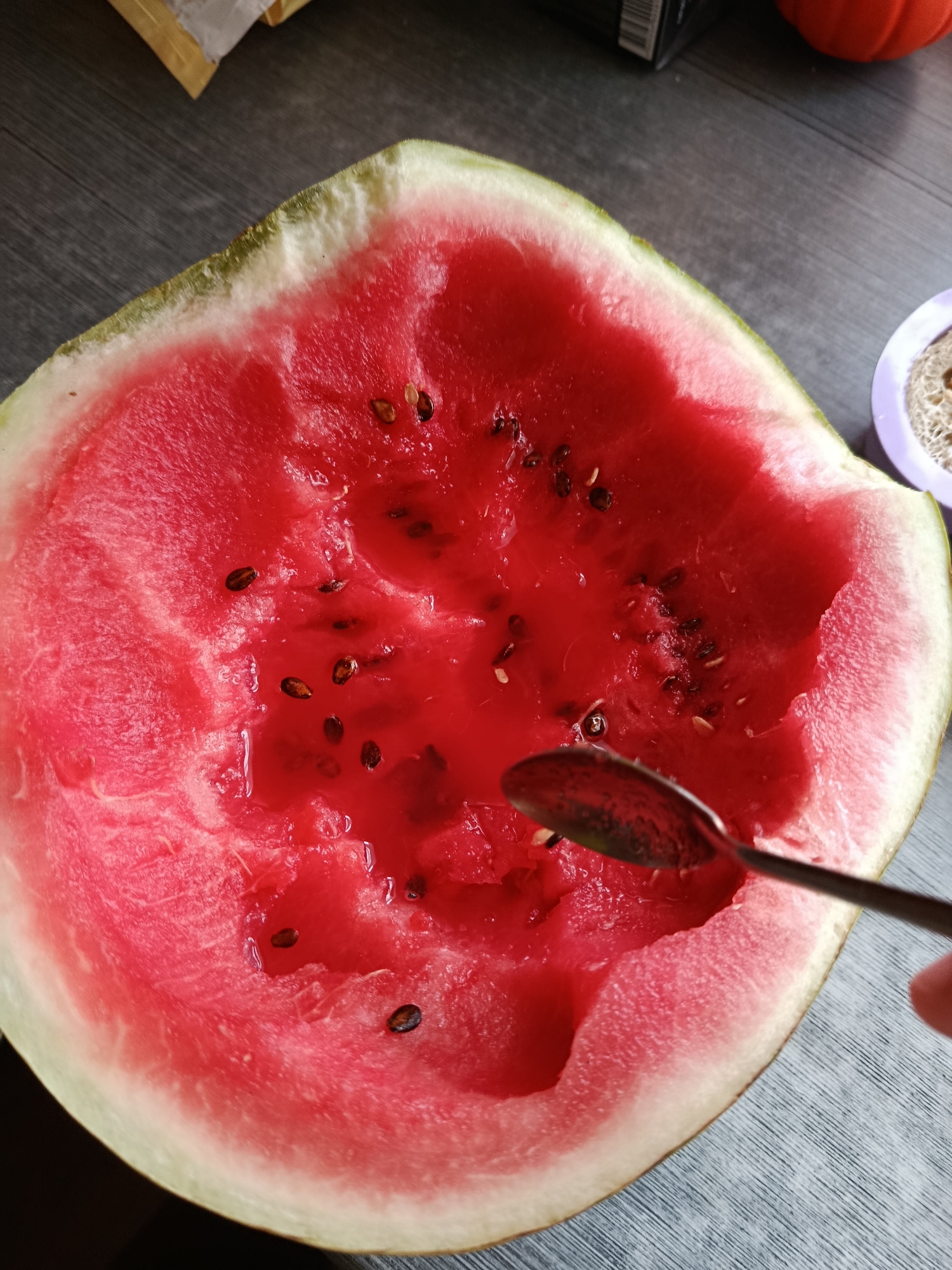 An overhead shot shows a halved watermelon on a dark wooden surface. A metal spoon is being used to scoop out the watermelon flesh from the rind. The watermelon flesh is bright red and has several dark seeds scattered throughout. The rind is a pale green and white. In the background, a purple dish with a sponge-like object is visible, as well as what appears to be packaging and a small orange object, possibly a decorative pumpkin. The scene is lit from above.