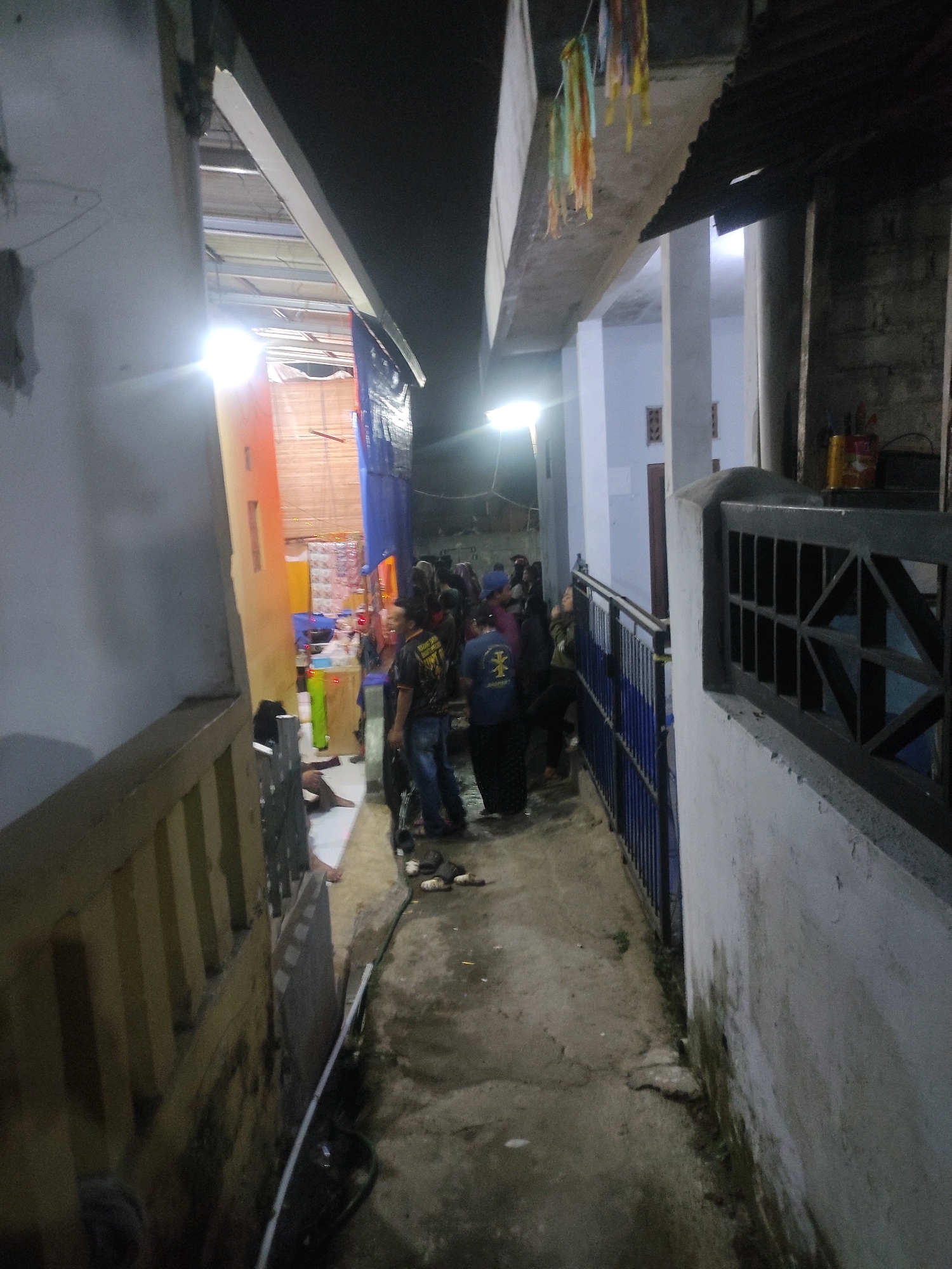 The image depicts a narrow outdoor alleyway at night, possibly in Padalarang, Indonesia. A group of people are gathered in the alley. Some individuals are standing, while others appear to be sitting or lying on the ground. The alleyway is bordered by buildings on either side. One side has a yellow wall with a railing and a dark-colored structure. The other side has a light-colored building with a blue metal fence. Overhead, there is a corrugated metal roof and some hanging decorations that appear to be streamers. The ground is paved with concrete and shows signs of wear and tear. Artificial lighting is present, casting a strong glow in some areas. The overall impression is of a casual gathering in a semi-urban or residential environment at night.