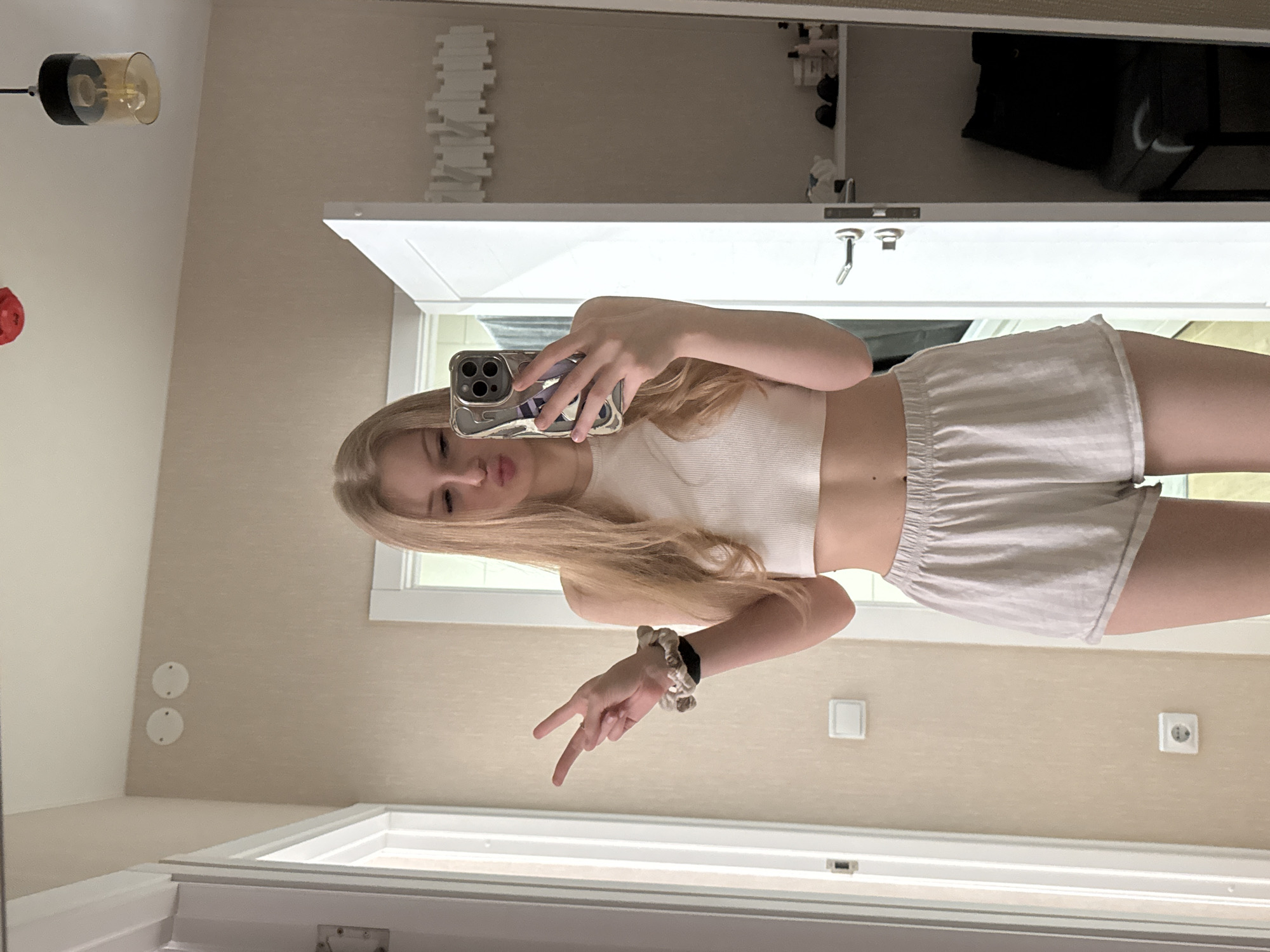 A young woman with long, blonde hair is taking a mirror selfie indoors. She is wearing a white crop top and light-colored shorts. She is holding her phone up with her left hand and making a peace sign with her right hand. The reflection shows a bathroom scene, including a vanity with a mirror, a light fixture, a wall with decorative elements, and electrical outlets. The location appears to be in a residential setting. The specific city, Perm, Russia, cannot be confirmed without further information.