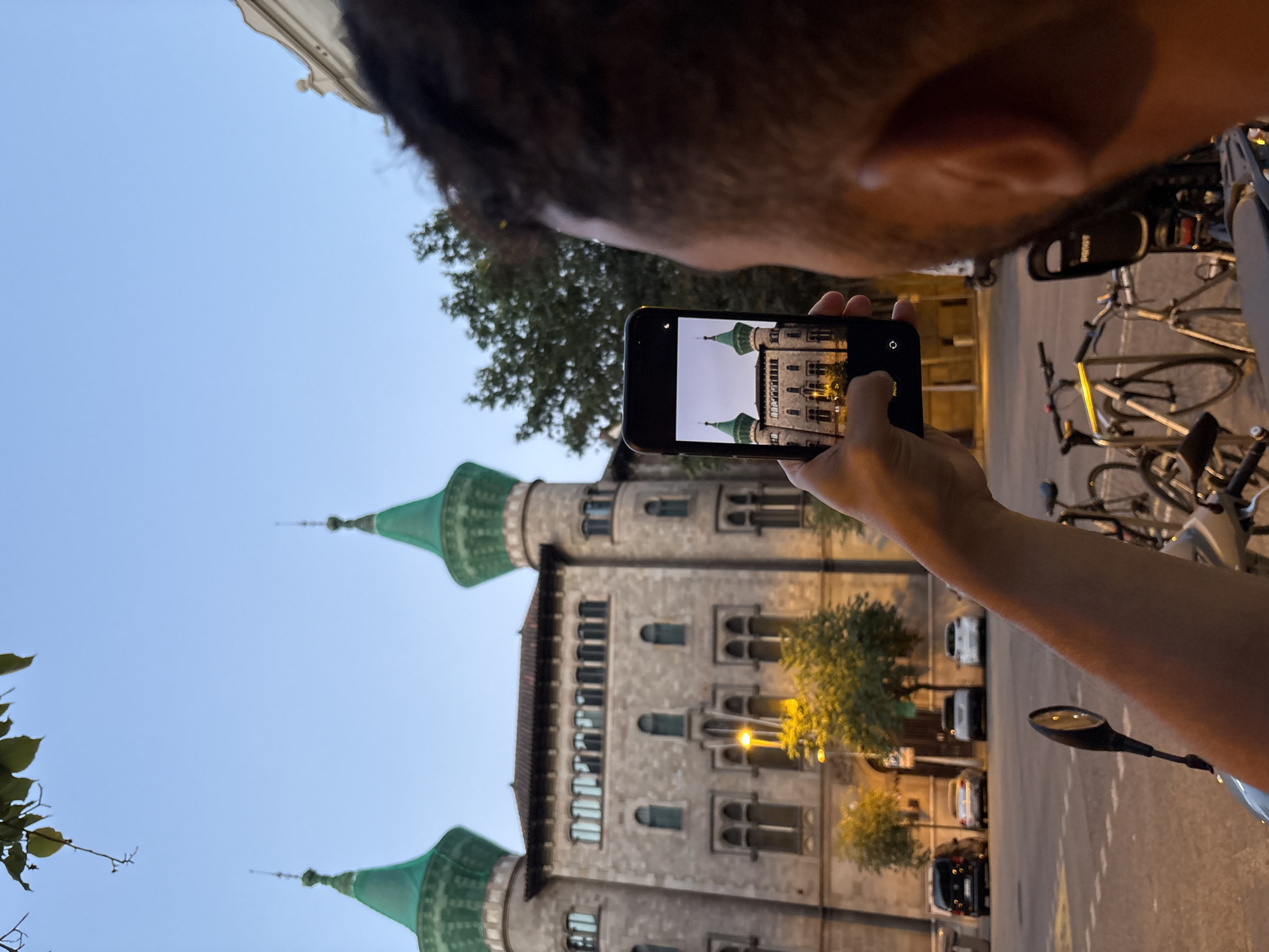 An over-the-shoulder shot shows a person holding a smartphone to take a picture of a building. The building is made of stone with two prominent green-capped towers, arched windows, and ornate architectural details. The smartphone screen displays a smaller image of the same building. In the foreground, a person's arm and shoulder are visible, along with parts of a scooter and bicycles. The background shows a street with parked cars, trees, and streetlights, suggesting an urban setting. The sky is clear and twilight, indicating the image was taken at dusk.
The building in the image is the Casa de les Punxes, located in Barcelona, Spain.