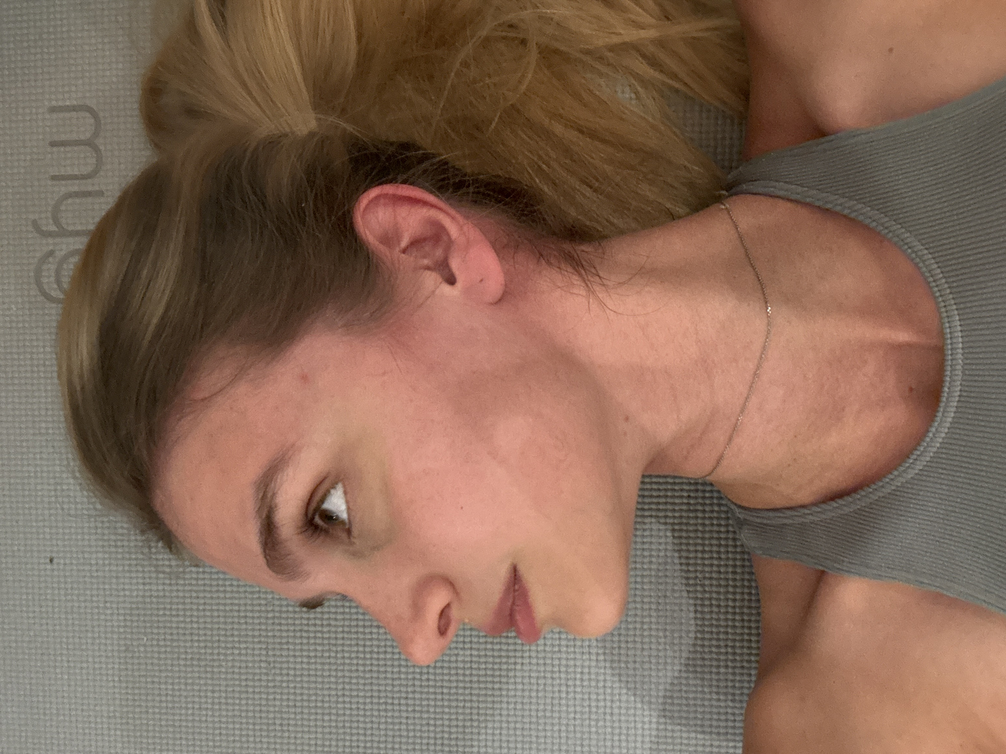 A fair-skinned woman with blonde hair pulled back into a ponytail is lying on a gray yoga mat with the brand name "myw" partially visible. Her head is turned to the left, and she is looking slightly upward. She has a gold necklace around her neck and is wearing a gray ribbed tank top. The background is out of focus and does not reveal any information about the location.