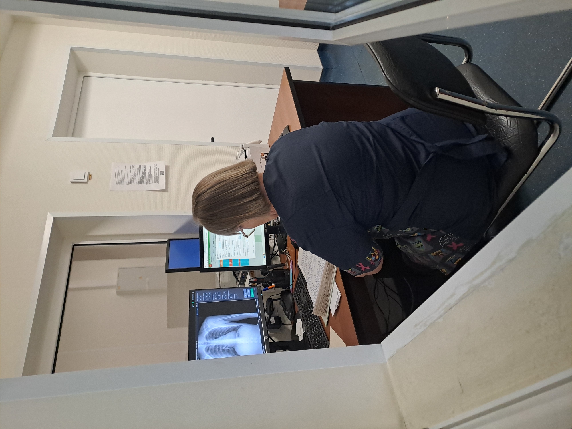 A medical professional sits in front of multiple computer screens displaying medical imaging, including an X-ray of a chest cavity. The individual is seated in an office environment, with paperwork and office supplies visible on the desk. Through an opening in the wall, a view of the office and hallway is visible. Without additional information, it is impossible to confirm the location in Elektrostal, Russia.