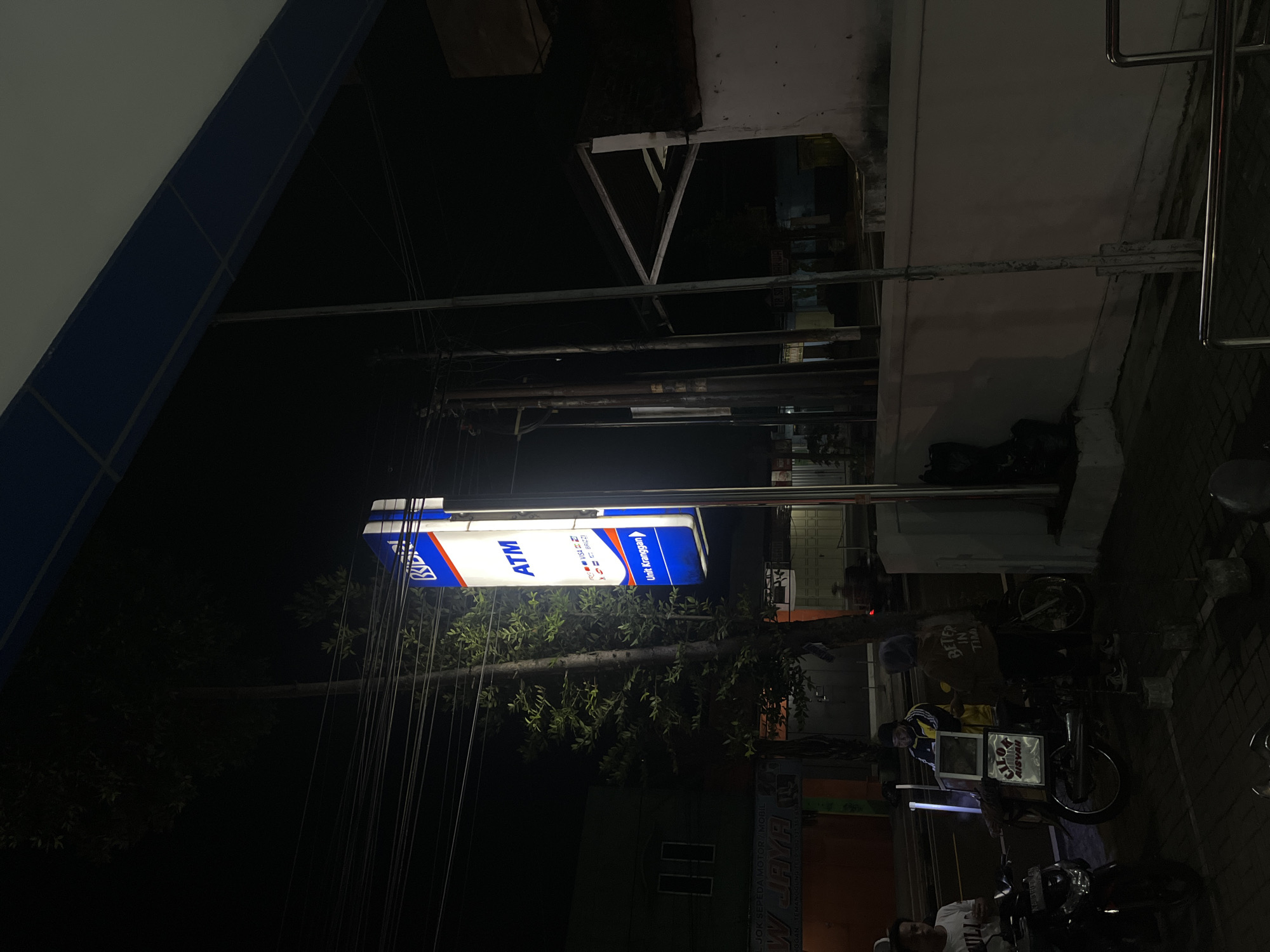 Here's a description of the image:
The photo was taken at night in Kranggan, Indonesia. The dominant feature is an illuminated ATM sign, advertising Visa and BRIZZI. There is a vendor selling food from a cart attached to a motorcycle, standing near the ATM. The vendor is dressed in a hat and shirt, and there are motorcycles nearby. The surrounding environment appears to be a street scene with foliage and buildings. Power lines are visible in the foreground. The image is oriented vertically.