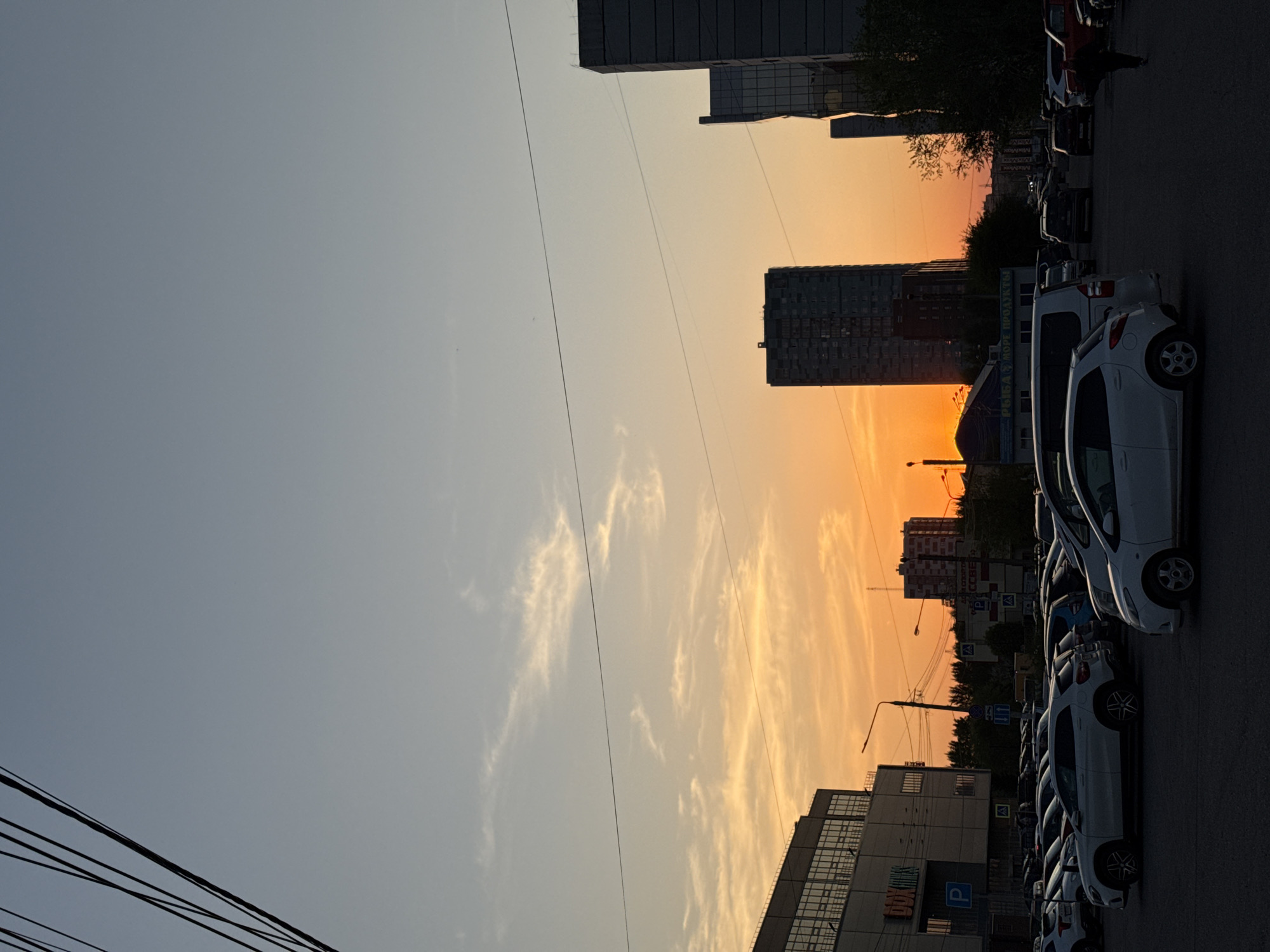 The image shows a street scene in Krasnoyarsk, Russia, at sunset. A row of parked cars lines the right side of the street, with several white cars visible in the foreground. Buildings of varying heights and styles form the backdrop, silhouetted against the orange and yellow sunset. Power lines crisscross the sky on the left side of the frame. The sky fades from light blue to orange near the horizon. The street and cars appear dark, emphasizing the bright sunset in the background.