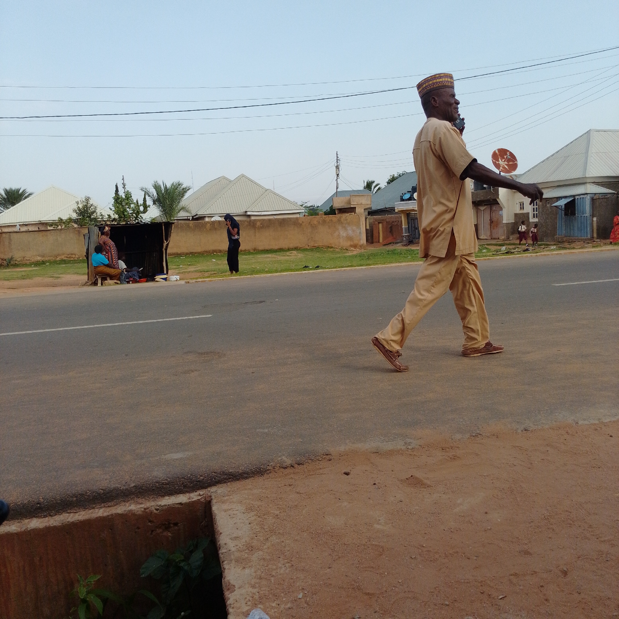 The image depicts a street scene in Gombe, Nigeria. In the foreground, a man wearing a light brown traditional outfit and a patterned cap walks across the asphalt road. He is looking to his left and appears to be speaking on a mobile phone. His right arm is extended outwards. In the middle ground, there is a makeshift stall with a corrugated metal roof, where several individuals are gathered. One woman in a blue top and patterned skirt sits on a stool, while another woman in a patterned dress stands. A person in a dark blue outfit is also visible near the stall. On the other side of the road, a person in a black garment walks away from the viewer. The background features houses with light-colored, sloped roofs, palm trees, and utility poles with overhead wires. The environment appears to be a moderately busy street with a mix of residential and commercial structures. The ground beside the road is covered in dirt and some sparse vegetation.