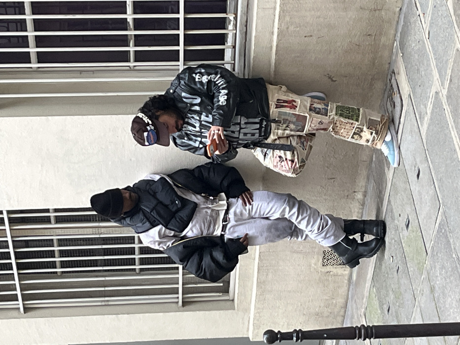 Two people stand next to a building. One wears a black jacket, light-colored pants, black boots, and a black beanie. The other wears a brown baseball cap, a black jacket with white writing, light-colored pants covered in images, and light blue shoes. Both face down, and the person with the baseball cap holds a small orange object. A metal railing runs along the bottom of the frame, and white horizontal grills appear behind both people.
