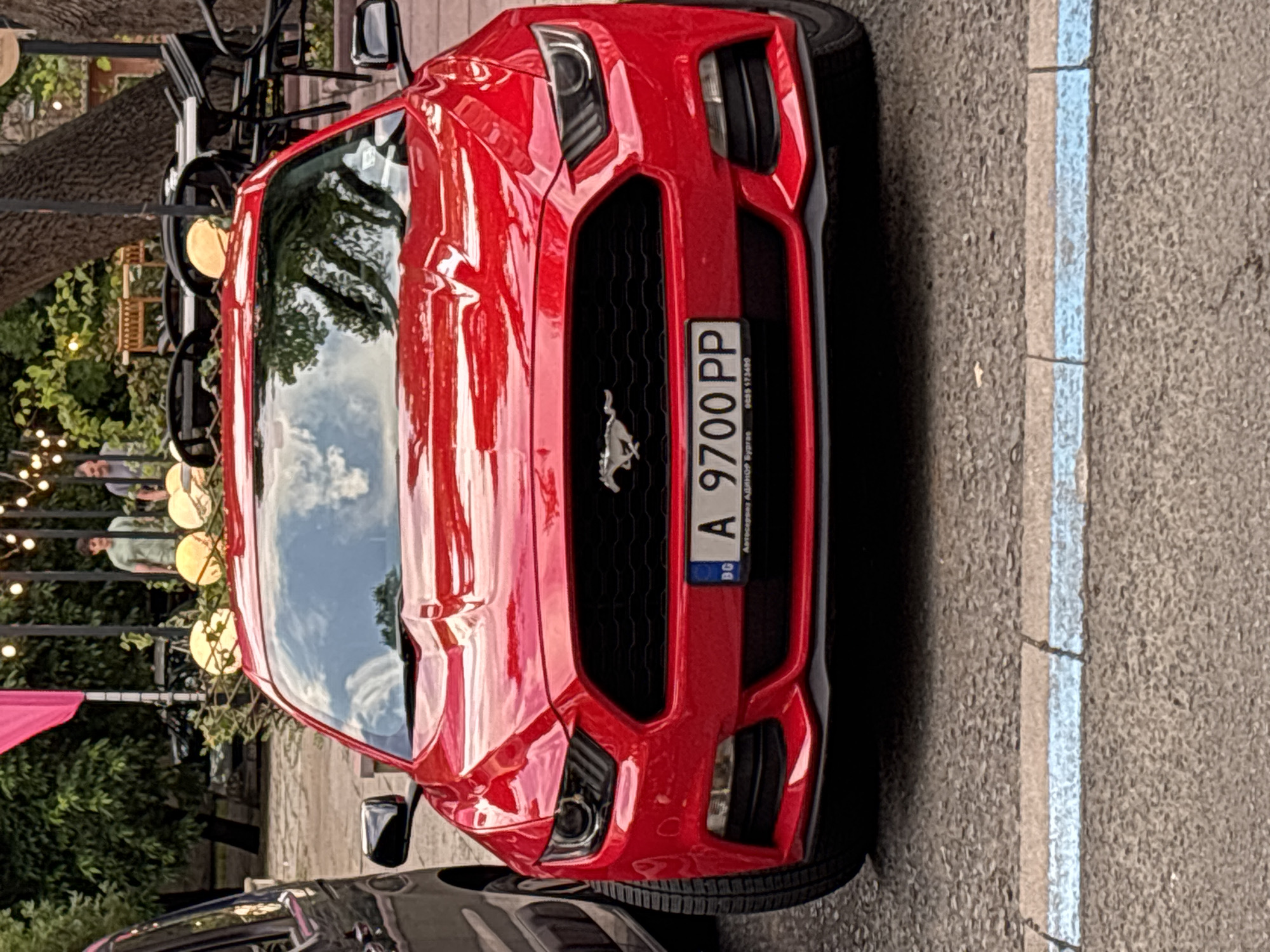 A bright red Ford Mustang is the central subject of this image, parked on a textured gray asphalt surface next to a light blue painted line. The car is viewed from a slightly elevated angle, showcasing its front fascia, including the distinctive pony emblem on the grille, aggressive headlights, and large air intakes in the bumper. The red paint is highly reflective, showing clear skies, white clouds, and green foliage from nearby trees.
A Bulgarian license plate is mounted on the front grille, displaying "A 9700 PP" and "BG". Below the main number, smaller text on the license plate frame reads "Автосервиз АДИНОРФ" (Autoservice ADINORF) and "0888 573499".
In the background, behind and to the side of the car, there appears to be an outdoor dining or cafe area. Several people are visible, some seated, others standing, reflected in the car's windshield and bodywork. One person in a light green shirt is clearly distinguishable. The area is adorned with decorative round, glowing lights or lanterns strung along what looks like an awning or trellis, and lush green trees provide additional cover. The scene suggests a pleasant daytime setting, likely in an urban or commercial area.
