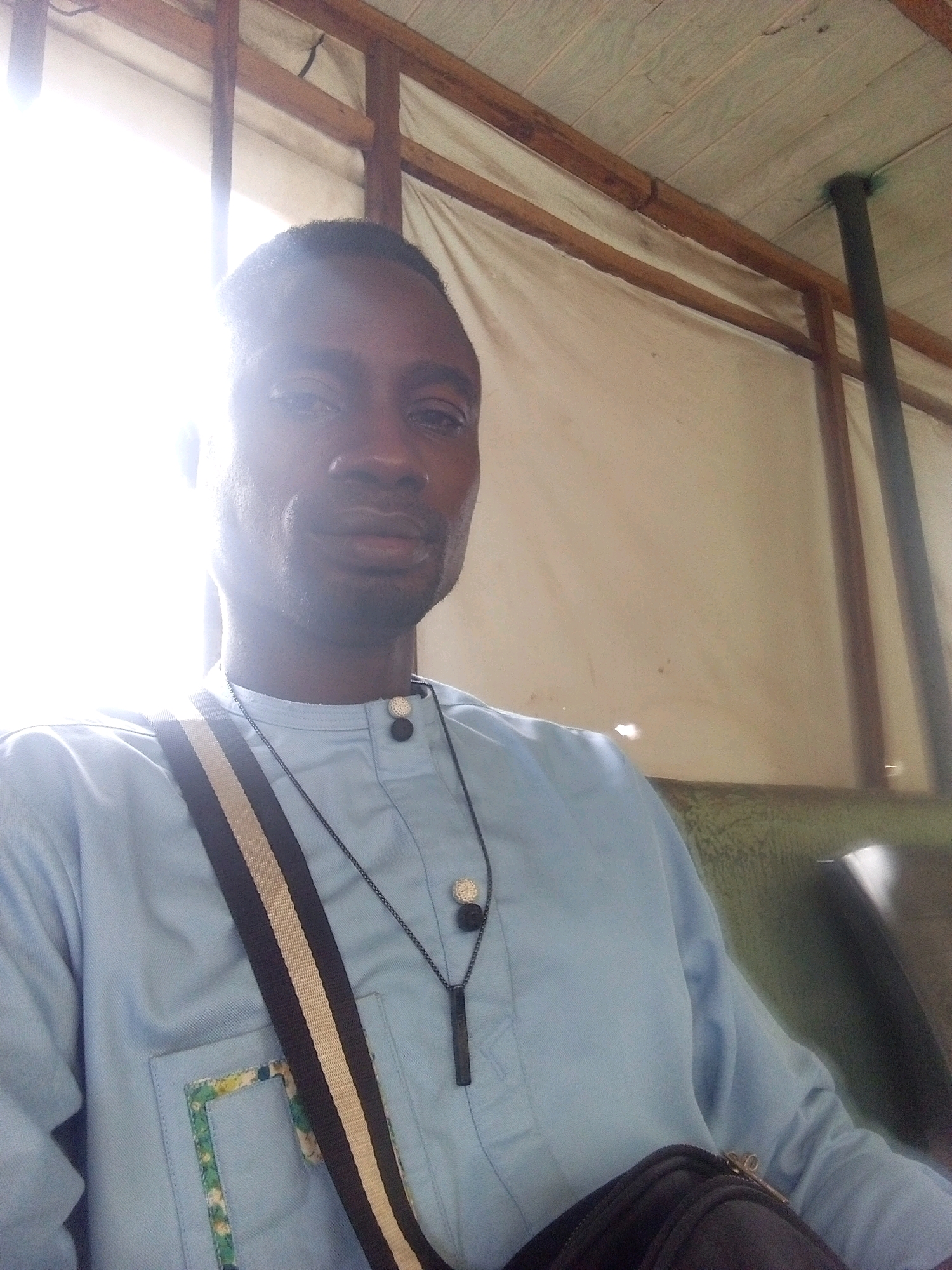 A medium shot shows a Black man with dark skin, short hair, and a light blue collared shirt with two black buttons. He wears a dark pendant necklace and a dark strap with beige and black stripes across his chest. He appears to be inside a structure with a ceiling made of wooden planks and beams, covered by a light-colored tarp. Sunlight streams in from the left, creating a bright glare. To the right, a dark pipe extends from the ceiling, and a dark green bench is visible. The man is looking forward, with his right shoulder closer to the camera. The setting appears to be in Zango-ndaji, Nigeria.