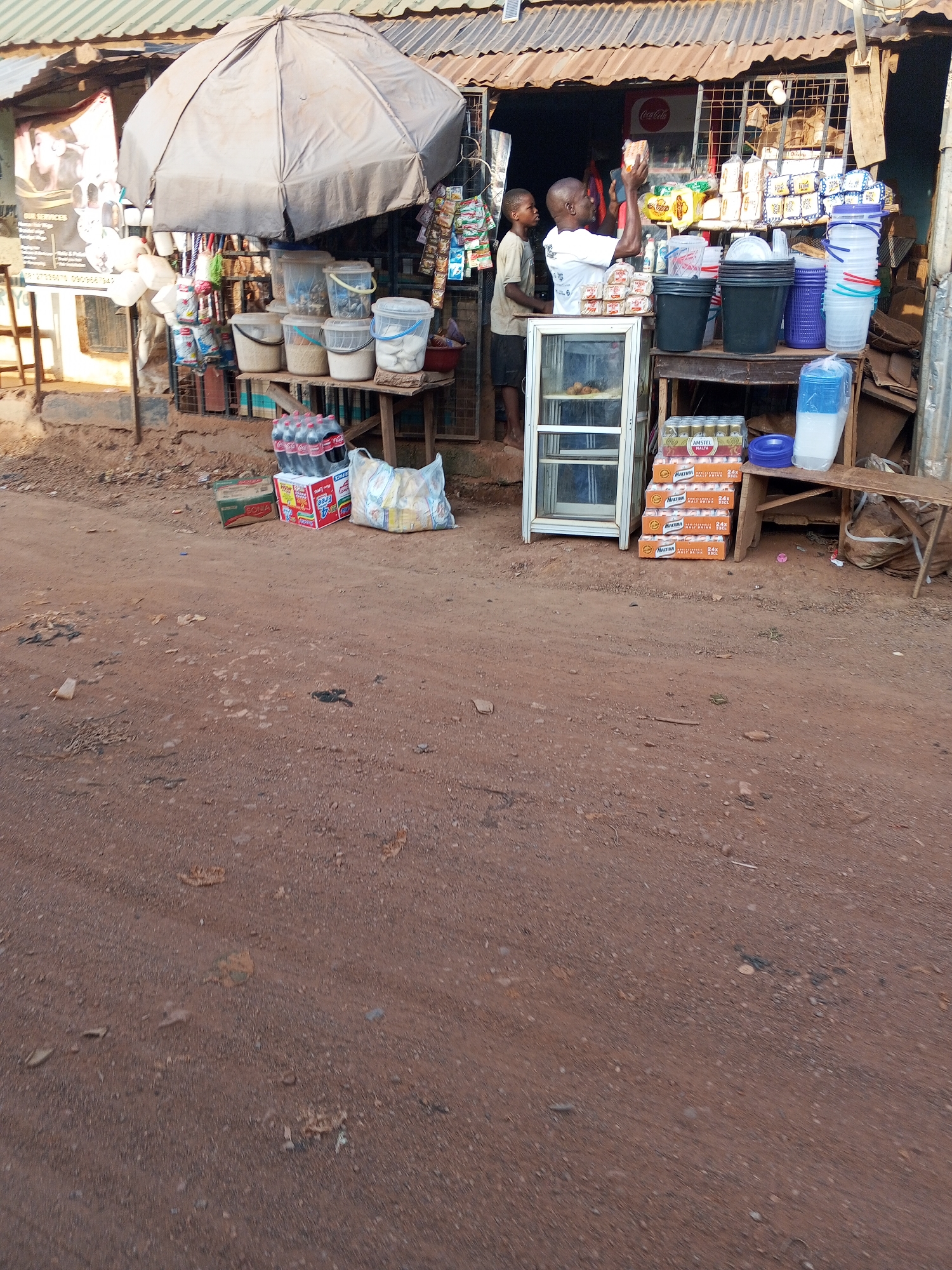 The image depicts a roadside retail shop in Abuja, Nigeria. The shop is characterized by a corrugated metal roof and wooden counters displaying a variety of goods, including beverages (Coca-Cola, Amstel Malta), household items (buckets, containers), and packaged snacks. Two male figures are visible within the shop's confines, one appearing to be attending to merchandise. A large, gray umbrella provides shade over a portion of the display area. The ground in front of the shop is unpaved and reddish-brown in color.