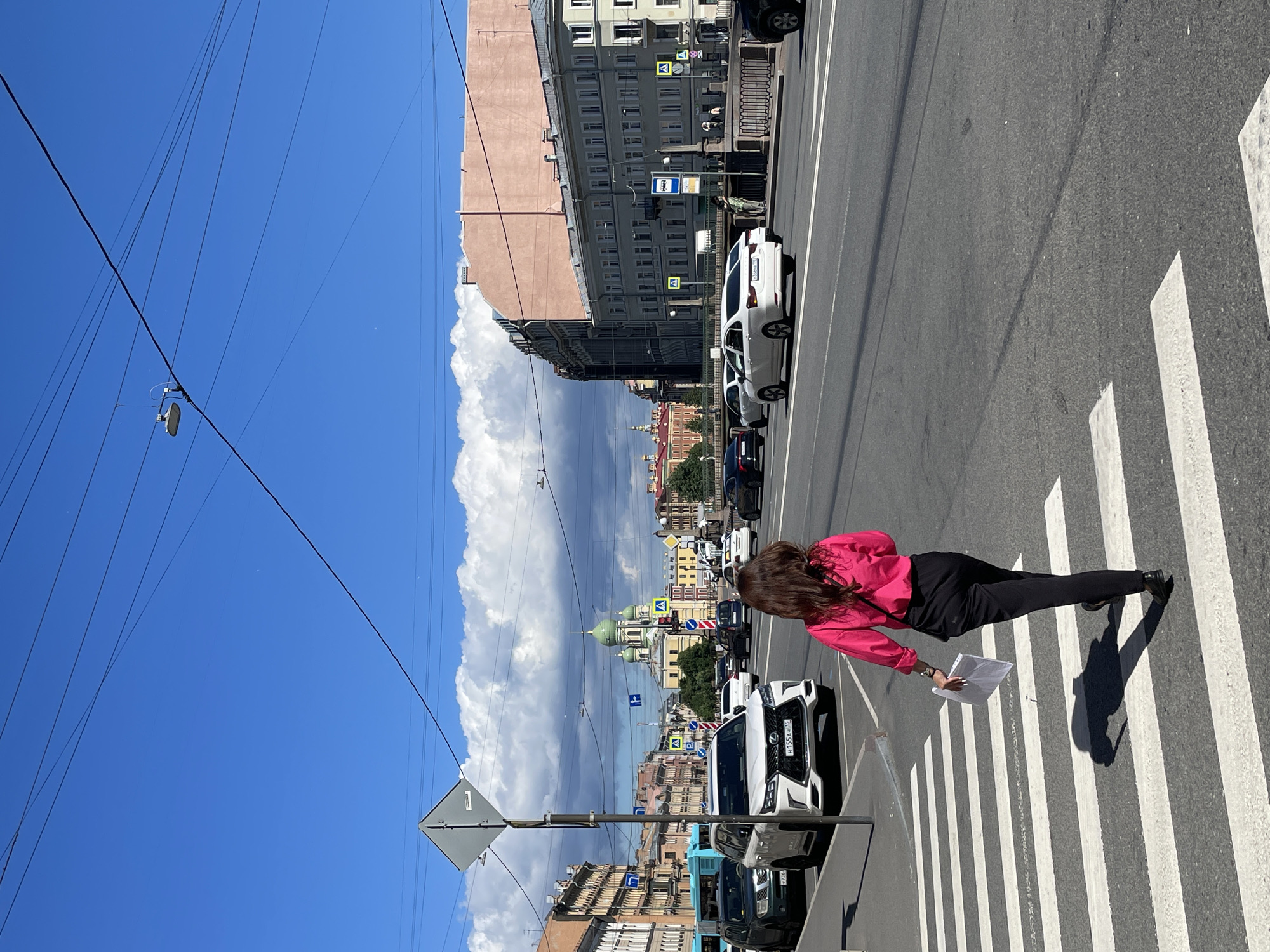 A woman walks across a zebra crossing on a wide, asphalt road in Saint Petersburg, Russia, during daylight. She is viewed from behind, wearing a bright pink collared shirt and black trousers. She carries a white document in her right hand. The sky is a clear, vibrant blue with large, white cumulus clouds. In the background, a green-domed church with a tall bell tower is visible, situated between buildings. The street is lined with multi-story buildings of various architectural styles, some with a lighter facade and others darker. Cars are present on the road, with a white Lexus SUV in the foreground to the left. Traffic lights and street signs are also visible. Overhead, a complex network of electrical wires crisscrosses the sky.