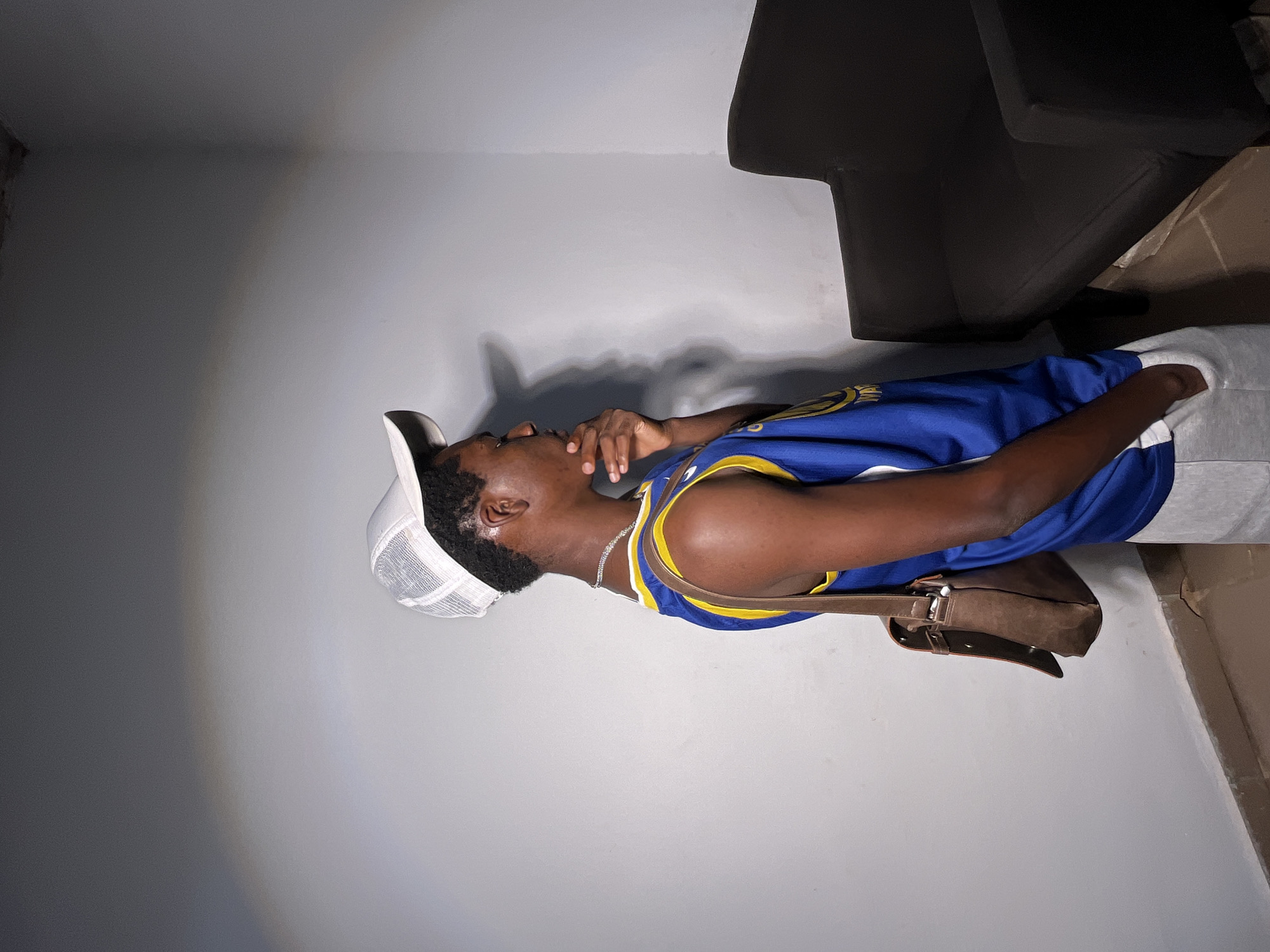 A side-profile, full-body shot shows a person wearing a white mesh baseball cap, a blue and yellow basketball jersey, and gray sweatpants. The person has dark skin and black hair. They are standing in an indoor setting against a light gray wall. A brown leather shoulder bag is slung across their chest, with the strap resting on their right shoulder and the bag itself hanging on their left side. The person's right hand is near their face, with fingers touching their chin, suggesting a pensive or contemplative mood. The lighting in the scene is dim, with shadows cast on the wall behind the person, indicating the light source is likely from the front and slightly to the side. The background includes a dark-colored armchair and a cardboard box. No visible text is present in the image. The time of day and weather are not discernible from the image.