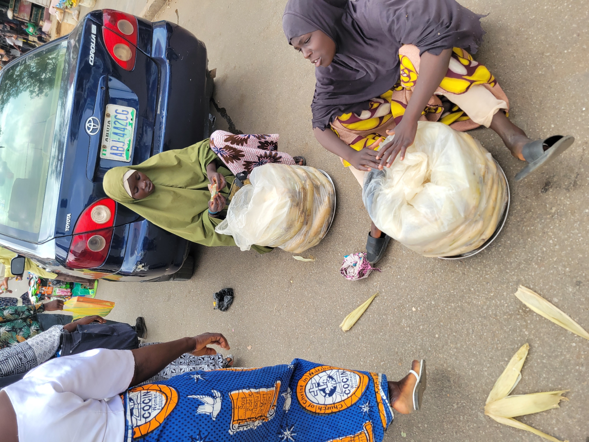 The image captures an outdoor scene, likely a street or roadside in Jos, Nigeria, given the license plate visible on the blue Toyota Corolla reads "ABJ-442CG" with "ABUJA" printed on it. Two women wearing hijabs and colorful clothing are seated on the ground next to the car's rear. They appear to be handling or selling what looks like bundled food items or produce wrapped in plastic, possibly corn on the cob, placed on metal trays. A third person, only partially visible, is also seated on the ground wearing a blue and orange patterned cloth, typical Nigerian attire, with a church-related logo. Corn husks are scattered around them. The environment is simple, with an asphalt surface.