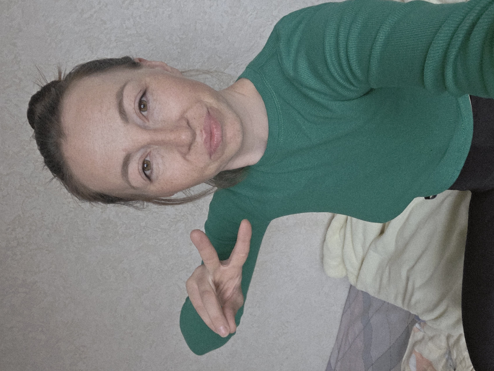 A woman is seen from a high-angle perspective, wearing a green long-sleeved shirt and black pants, against a backdrop of an off-white wall and a bed with neutral-colored sheets and a patterned blanket. She has her hair pulled back in a bun, and she is making a peace sign with her right hand. The location is a bedroom in an unknown city.
