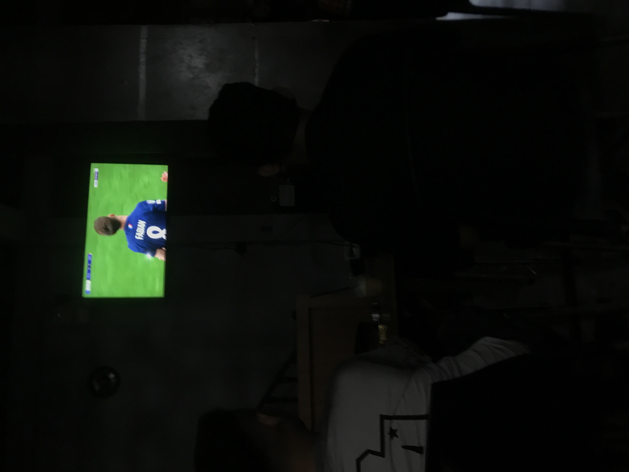 In a dimly lit room in Lhokseumawe, Indonesia, a person wearing a white t-shirt with a black design is partially visible in the foreground. A television screen displays a football video game, showing a player with the name "FABIAN" and the number 8 on his blue jersey. Another person is partially visible, obscured by darkness. The room is poorly illuminated, making it difficult to discern details.