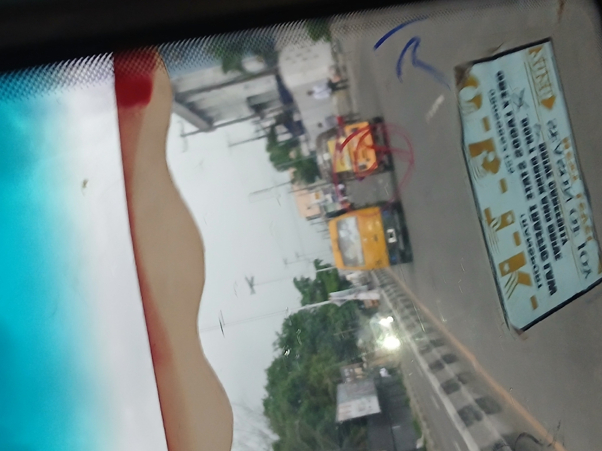 The image appears to be taken from inside a vehicle, possibly a car or bus, looking out through the windshield at a street scene in Animashawun, Nigeria. The view is somewhat obstructed by the vehicle's interior, including the blue tinted windshield and the side pillar on the left.
Visible through the windshield are buildings, trees, and vehicles on the road, including yellow buses, a car, and what appears to be a smaller motorized vehicle. There is also a street sign or notice taped to the windshield, obscuring some of the view and bearing text. The road appears to be paved. The overall environment suggests an urban setting.