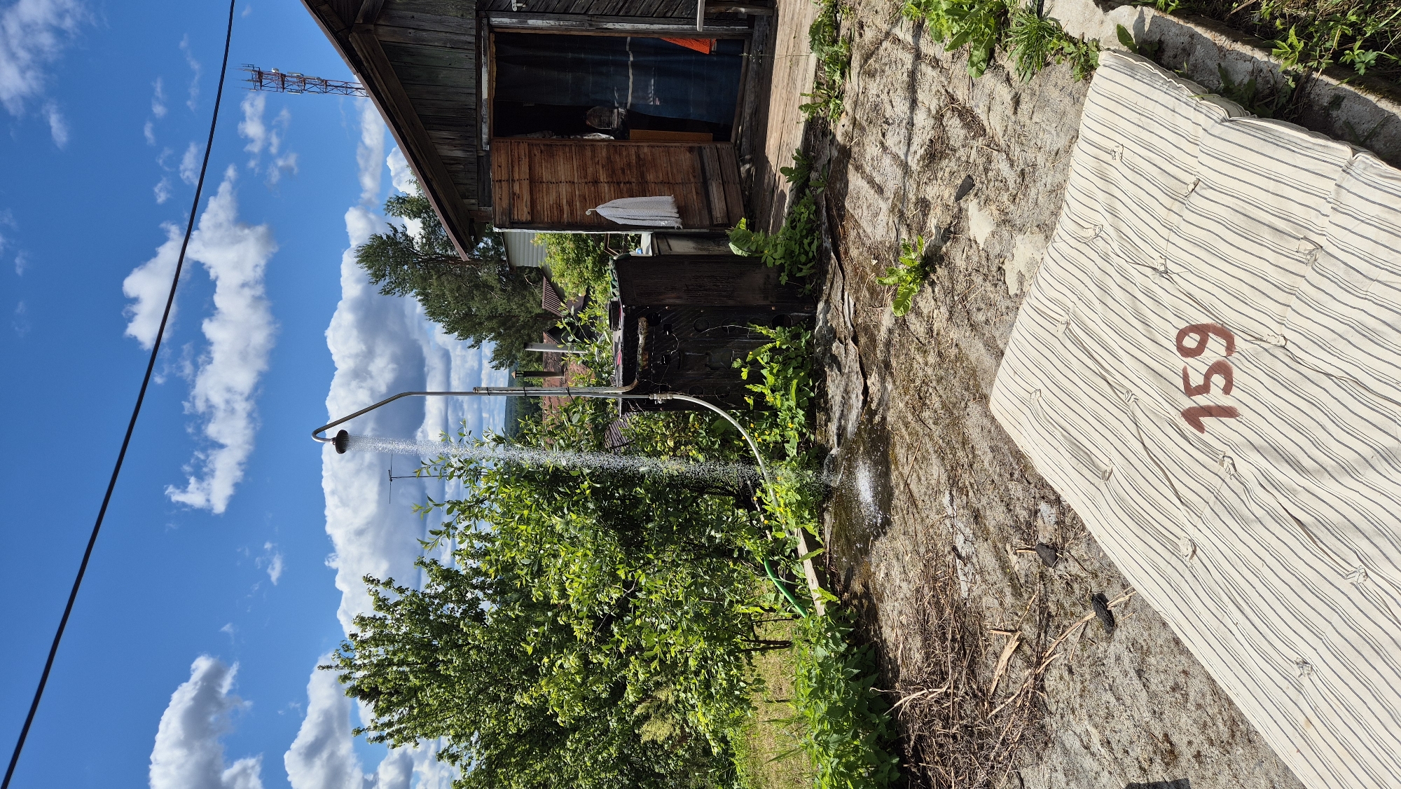 The image depicts an outdoor shower setup next to a rustic wooden structure. Water is actively spraying from a showerhead attached to a curved metal pipe. The showerhead is positioned over a dark, rectangular tank, which appears to be part of the shower system, possibly a water heater or storage. A hose snakes from the tank to the showerhead. Green vegetation, including bushes and trees, surrounds the shower area and the wooden building. The building has an open doorway revealing a blue curtain inside and a white towel hanging on the exterior. In the foreground, a striped mattress with the number "159" stenciled in red is placed on a concrete or stone patio. The ground is a mix of concrete, dirt, and dry debris. The sky above is blue with scattered white clouds, and a power line stretches across the top of the frame. A cell tower is visible in the distance. The scene is set in a rural environment, consistent with a location in Машезеро, Russia, suggesting a simple, utilitarian setup for outdoor living or bathing.