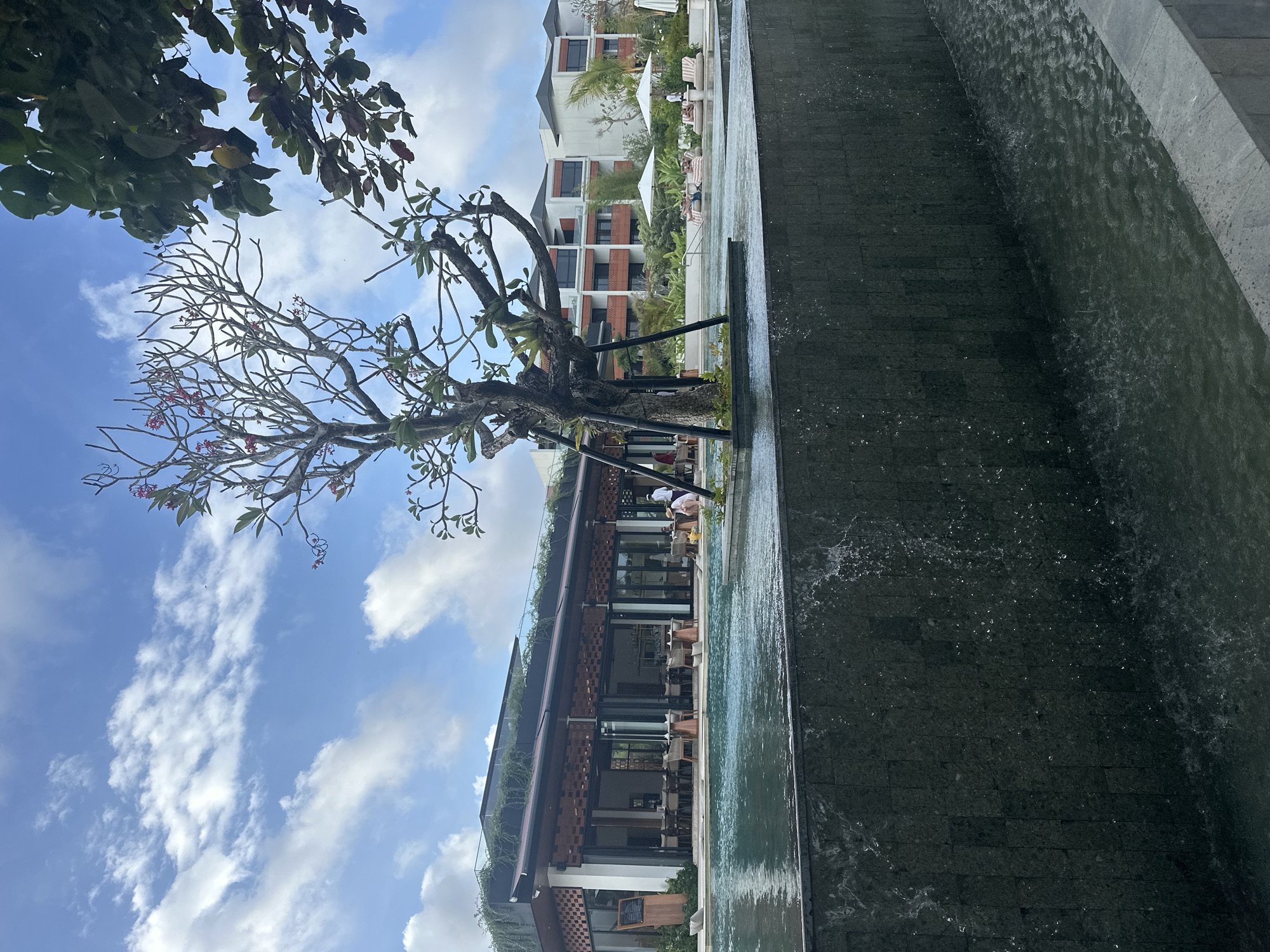The image depicts an outdoor scene in what appears to be a resort or upscale hotel, potentially located in Denpasar, Indonesia, based on the architectural style and vegetation. A dark stone waterfall feature runs along the right side of the frame. To the left, a tree with sparse foliage stretches towards a blue sky with scattered clouds. A multi-story building with a restaurant or dining area is situated behind the tree. White lounge chairs and umbrellas are visible in the background, suggesting a pool or relaxation area. People are present but not distinctly visible.