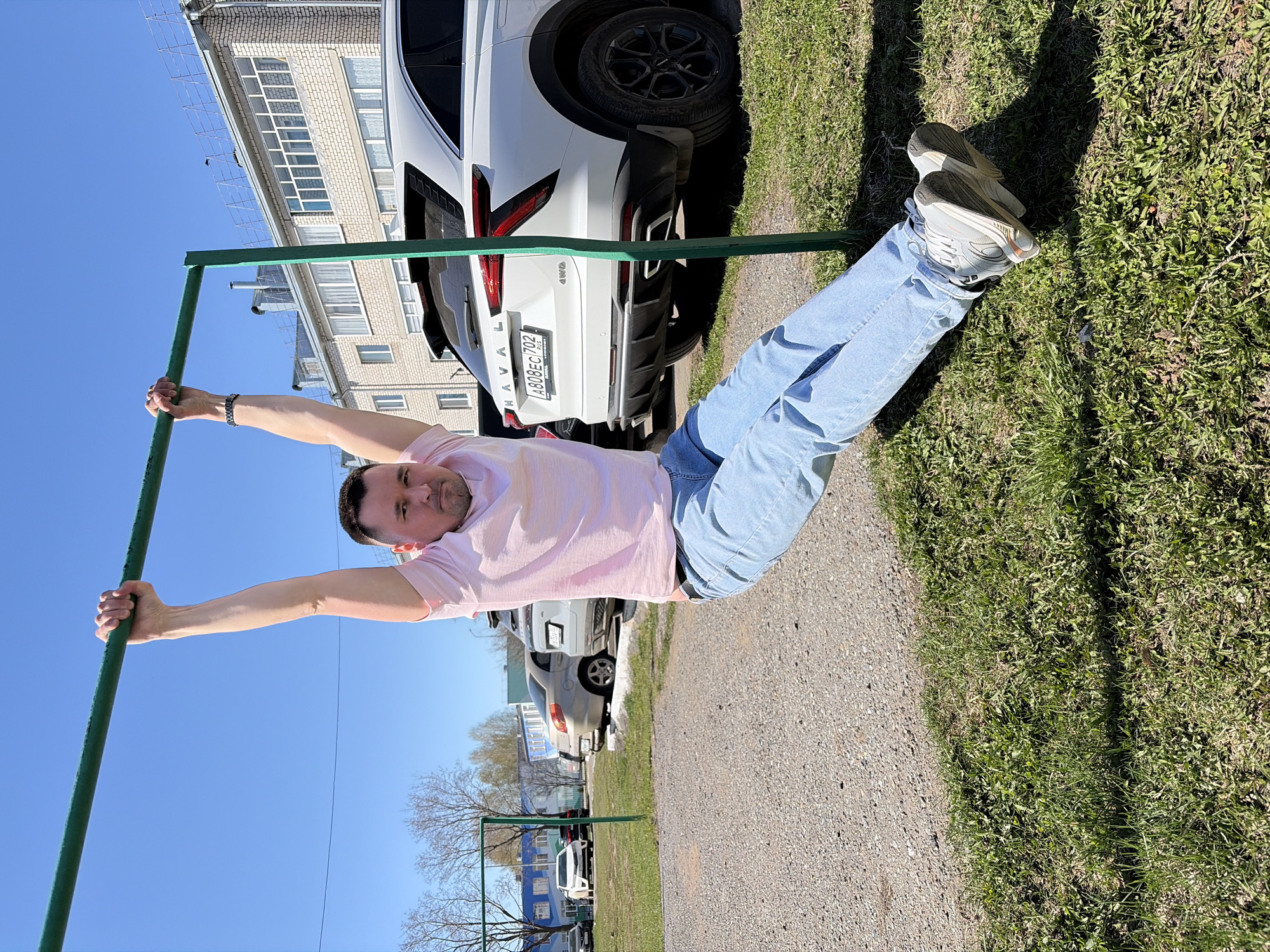 A man in light blue jeans and a pink shirt hangs from a green metal bar outdoors. The man is facing the viewer. His arms are extended, holding the bar, and his legs are straight, angled toward the grassy ground. He is wearing grey sneakers. Behind him is a white Haval car and the corner of a brick building. Further in the background are more parked cars and trees with bare branches. The sky is a bright blue. The location is Neftekamsk, Russia. A swing set is visible behind the man.