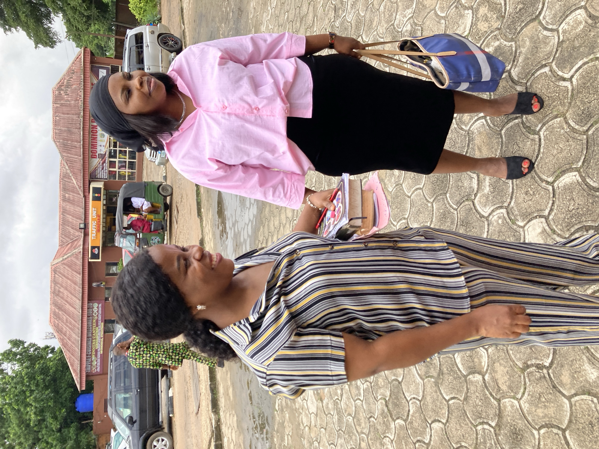 Two Nigerian women are standing on a patterned paved surface outdoors in Aba, Nigeria. The woman on the left is wearing a black, white, and yellow striped shirt and matching striped pants. She is smiling and holding a stack of books and pencils. The woman on the right is wearing a pink button-down shirt, a black skirt, and a black headscarf. She is also smiling and holding a blue and white striped tote bag. In the background, there are buildings, cars, and trees. One building has signs that read "TRIPLE F CUISINES WELCOMES YOU TO WINNERS CANTEEN" and "TRAFFIC UNIT". Another building has a sign that reads "DOMINION". The ground appears wet in some areas, suggesting recent rain.