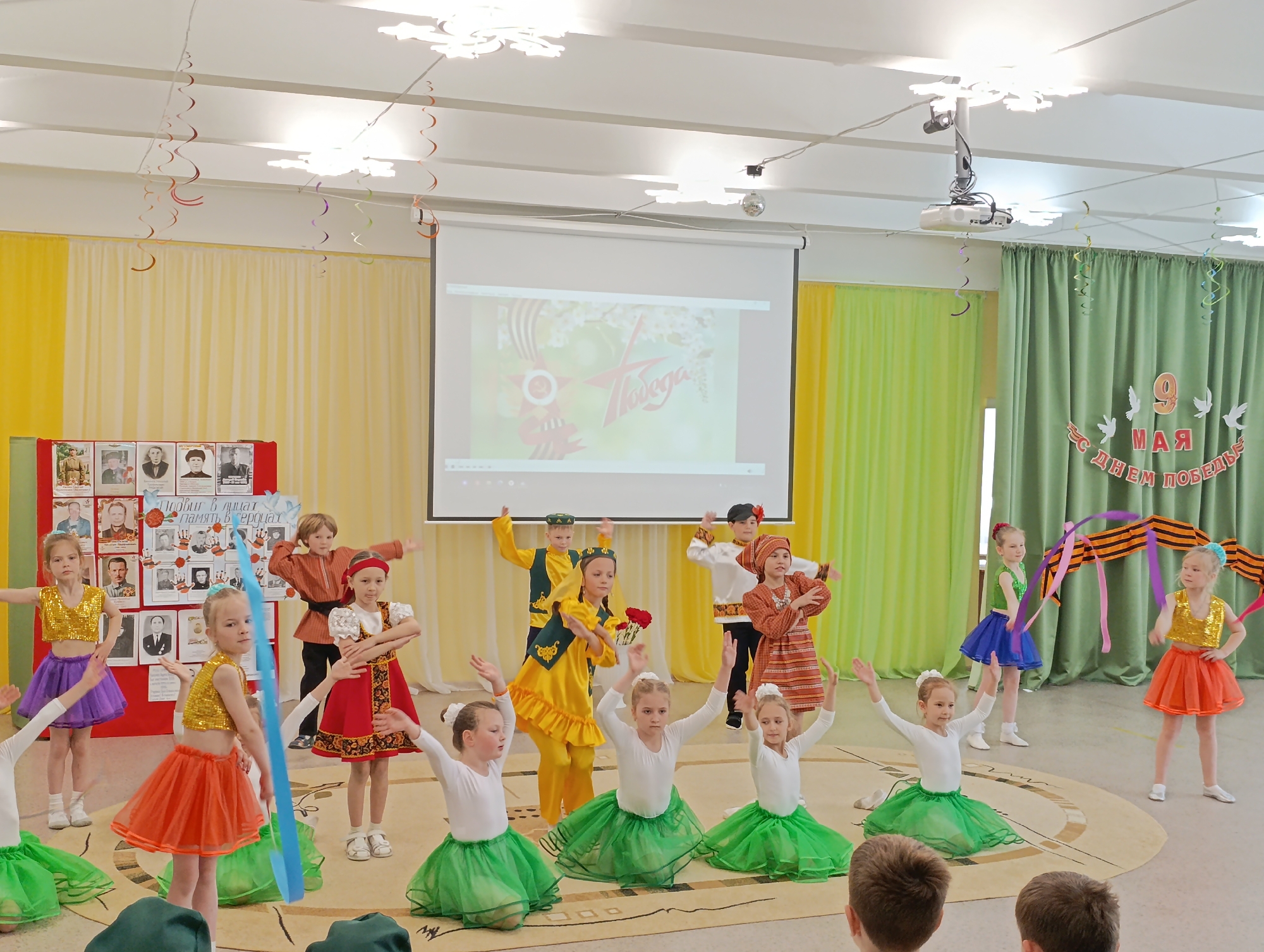 A group of children are performing on a stage in a room decorated for Victory Day (May 9) in Chaikovsky, Russia. There are approximately 15 children dressed in various costumes, including green tutus with white tops, gold sequined tops with colorful skirts, and traditional Russian clothing. Behind them is a projection screen displaying a Victory Day theme. To the left of the screen is a display with images of individuals, possibly war heroes. To the right of the screen is a backdrop with a "9 May" banner decorated with doves and a St. George ribbon. Decorations hang from the ceiling. The room appears to be a school hall or auditorium. Adults are likely present in the audience.