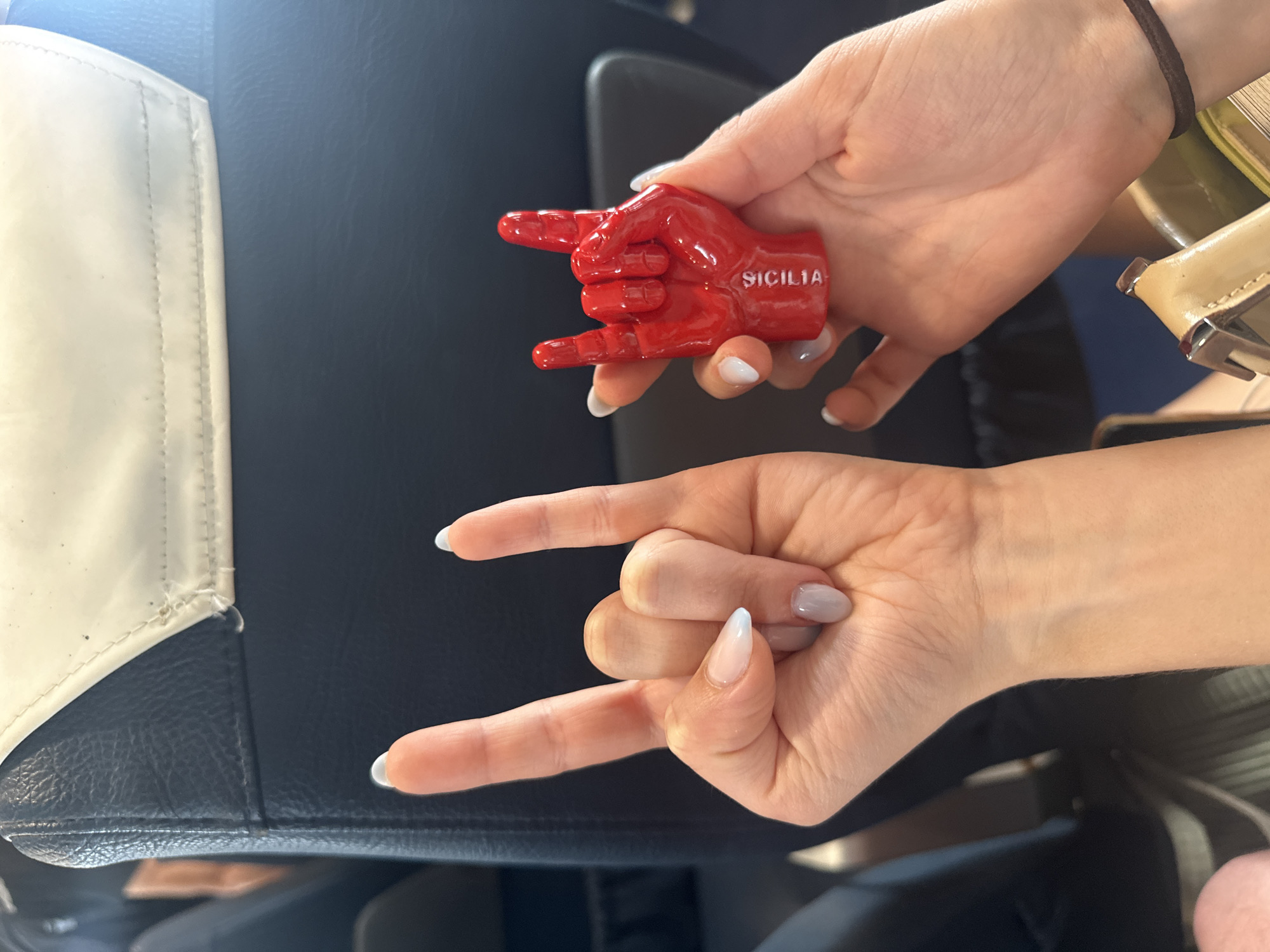 Two hands are prominently featured, one holding a red, glossy souvenir and the other making a hand gesture, set against a dark blue or black upholstered background, likely a vehicle seat with a white trim.
One hand, with light pink and white ombré nail polish, grasps a bright red, hand-shaped object forming the "rock on" or "devil horns" gesture. The word "SICILIA" is embossed in white letters on the side of this object, indicating its origin or connection to Sicily. The other hand, with light grey/lavender nail polish, mirrors the same "rock on" gesture. A dark brown hair tie is visible on the wrist of the hand holding the souvenir, and a light brown bag or purse is partially seen in the background.
The scene suggests a lighthearted moment, possibly a celebration or a memento from a trip. The "SICILIA" text directly links the item to Sicily, making the location context of Milazzo, Italy, highly relevant. The setting appears to be indoors or within a mode of transport like a car or plane.