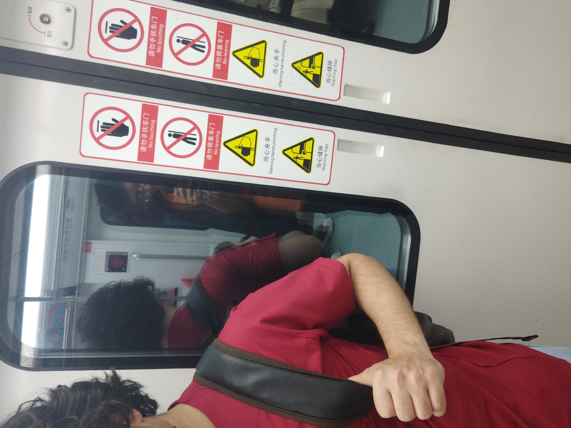 The image shows a person with dark, curly hair and a red shirt standing inside a train car. The person is standing near a window and is reflected in the glass. The door has signs that read "No touching," "No leaning," "Warning hands pinching," and "Warning Gap" in both Chinese and English. The location in the city is Unknown, Unknown.