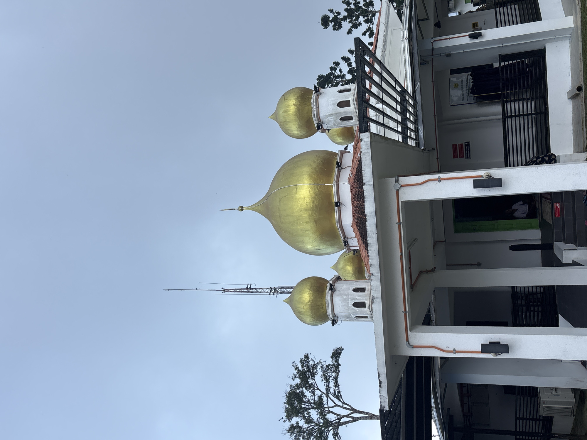 This is a medium shot, eye-level, outdoor photograph of a mosque with golden domes under a cloudy sky. The mosque has a white exterior with orange piping visible on the outside of the building, and is partially shaded by a black metal awning structure. Multiple golden domes of varying sizes are visible on the roof of the building, with a tall antenna rising behind the largest dome. A mature tree with dark green foliage is located to the left of the mosque. The foreground shows a white, covered walkway leading to a green doorway, with a person visible inside. The image appears to be taken in George Town, Malaysia.