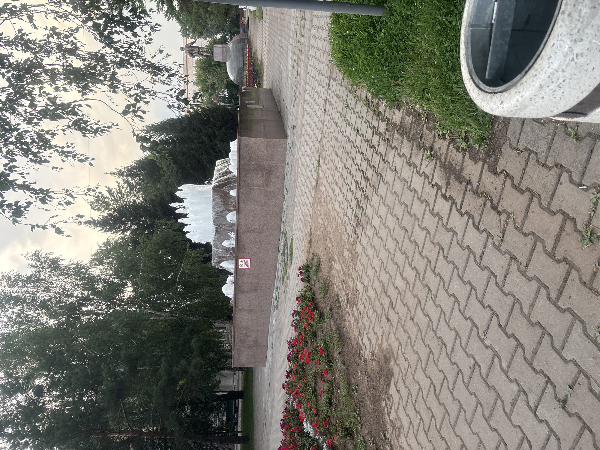 The image depicts a public park scene in Pavlodar, Kazakhstan, featuring a large stone fountain with water jets spraying upwards. Surrounding the fountain is a plaza paved with interlocking concrete blocks, with a flowerbed of red and pink flowers in the foreground. In the background, there are various trees, including conifers and deciduous varieties, as well as a statue on a pedestal and a building. The sky is overcast, suggesting a cloudy day. The overall impression is that of a well-maintained urban green space.