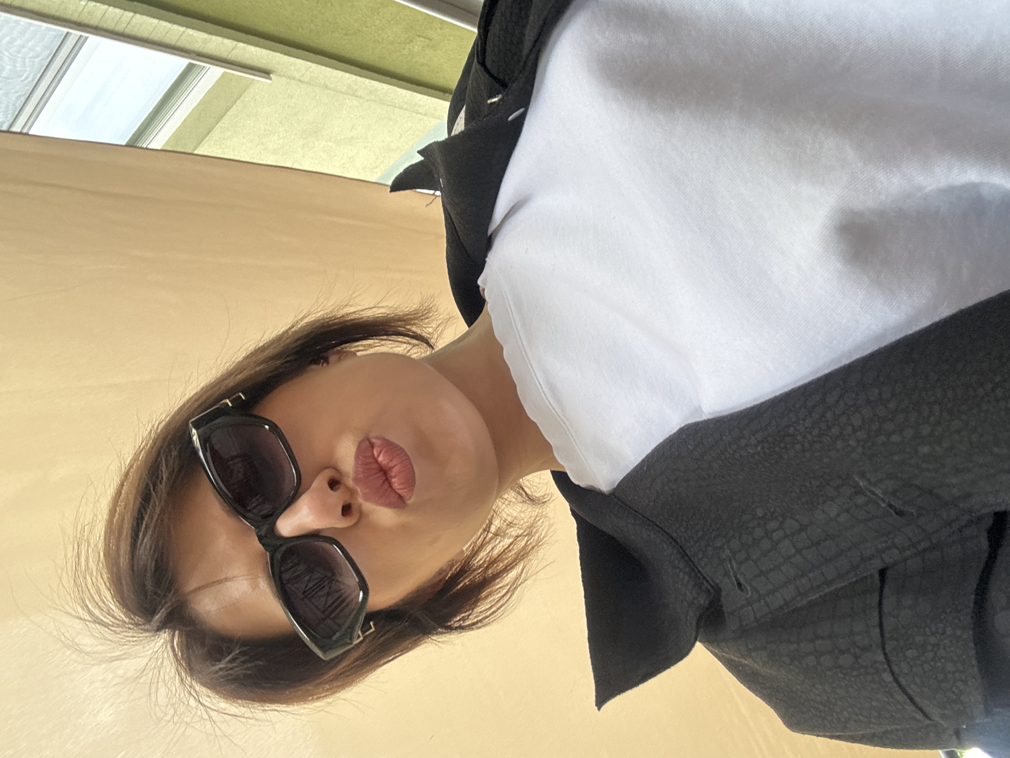 A person is looking down at the camera, wearing sunglasses, a white shirt, and a black jacket. The location appears to be outdoors, possibly in Sofia, Bulgaria, based on building architectural elements. The background includes a beige-colored wall and a window with green trim. The person is making a kissing face.