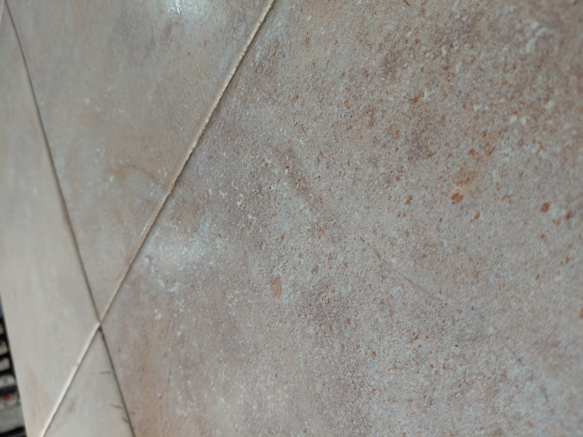 A close-up, angled shot captures a tiled surface, likely a floor or wall, with a light beige and brown mottled pattern. The tiles are arranged in a grid, separated by narrow grout lines. The surface of the tiles exhibits a granular or speckled texture with irregular lighter and darker patches, resembling natural stone. Some areas show a slight sheen, suggesting a polished finish or the reflection of ambient light. The overall color palette is warm, dominated by various shades of light brown, cream, and off-white, with small, darker brown speckles scattered across the surface. No people or specific objects other than the tiles are visible. The setting appears to be an interior space. There are no discernible location cues within the image itself to confirm the Santa Ana, Venezuela context. No text is visible.