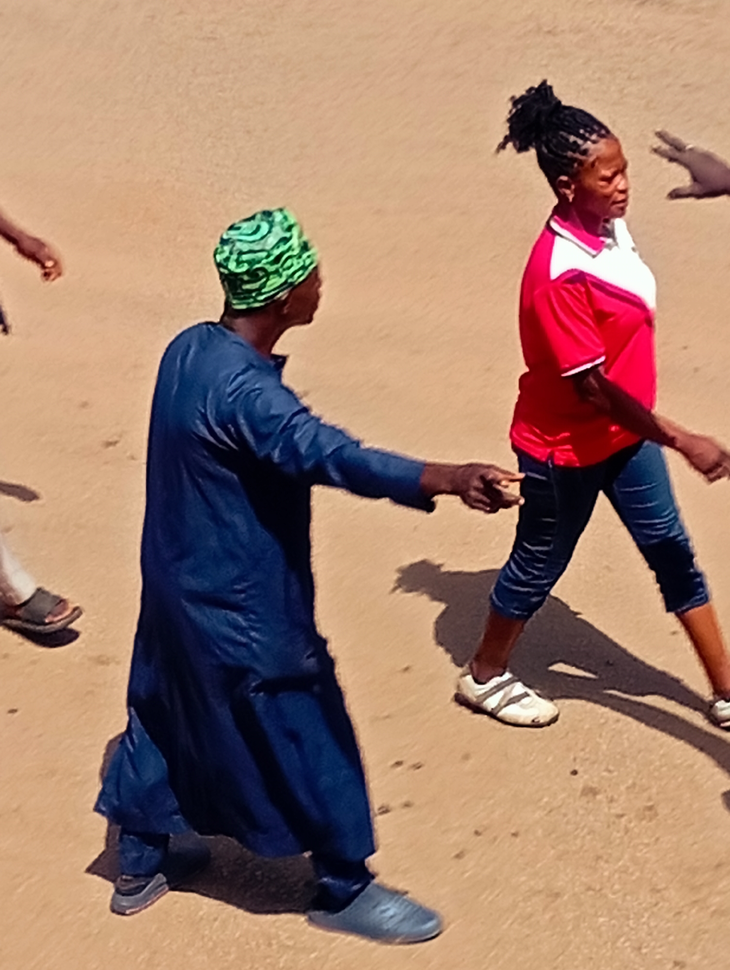 In Bukuru, Nigeria, a group of people are walking on a sandy, unpaved surface. In the foreground, a person wearing a blue garment and a green and black headwrap is walking towards the right. Their right hand is extended, and they appear to be interacting with someone ahead. To their right, a woman with braided hair tied up in a bun is walking in the same direction. She is wearing a red and white collared shirt and dark-colored cropped pants, along with white athletic shoes. Further to the right, another person's hand is visible in the frame, reaching towards the woman. In the background, to the left, another individual's legs and feet are visible, wearing dark sandals. The ground is a uniform light brown, suggesting an outdoor, possibly dry environment. Shadows are cast on the ground, indicating bright sunlight.