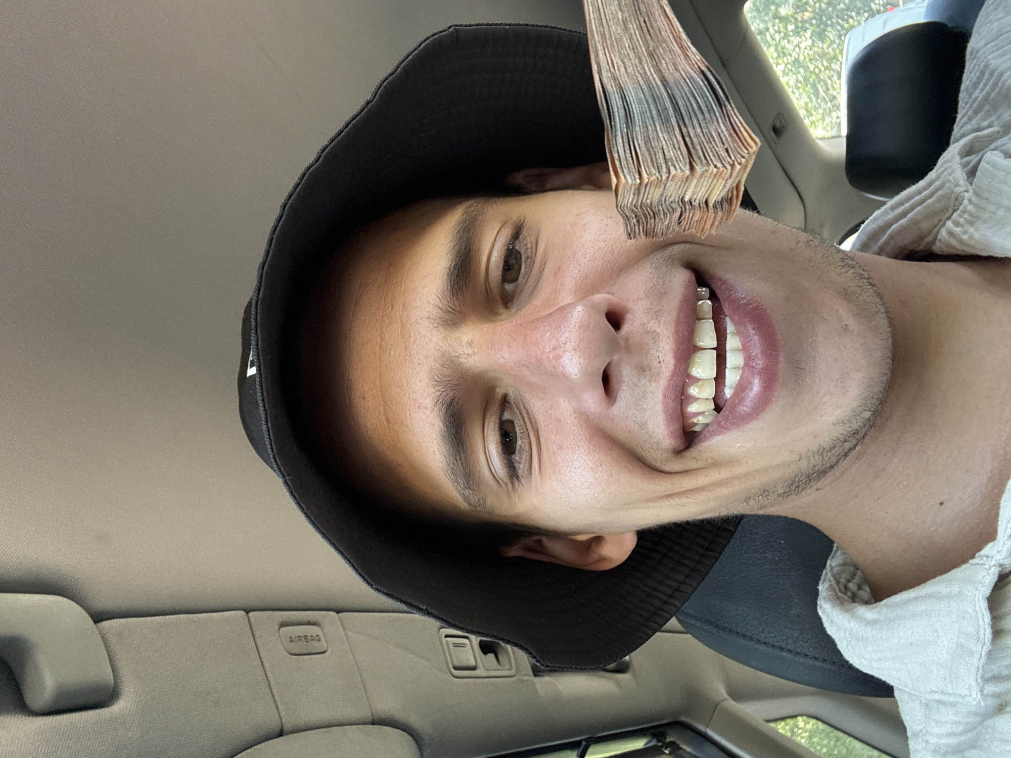 A close-up, front-facing, selfie-style image captured inside a vehicle shows a young man wearing a black bucket hat and a light-colored, textured shirt. He is smiling broadly, revealing his teeth, some of which have visible dental work, potentially grills. His right hand is holding a thick stack of banknotes in front of his face, obscuring part of his lower facial features. The background of the image reveals the interior of a car, including the gray headliner with an "AIRBAG" sticker visible on the passenger side, and a dark car seat. Through the vehicle's windows, glimpses of greenery and sunlight suggest an outdoor setting, possibly within the city of Sofia, Bulgaria, given the user's prompt. The lighting is natural and somewhat harsh, casting shadows on the man's face.