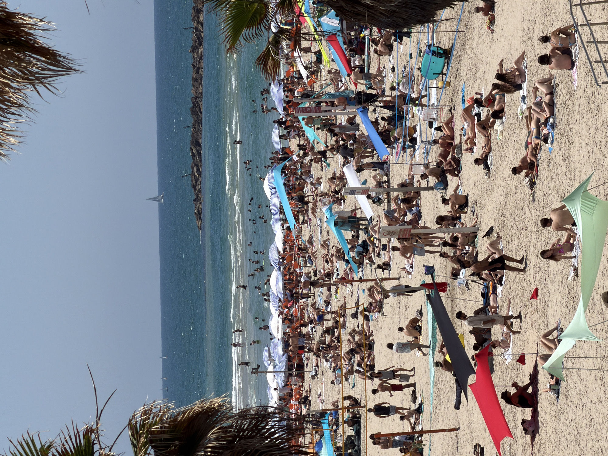 The image shows a crowded beach in Tel Aviv, Israel. People are sunbathing, swimming, and walking along the shore. There are numerous beach umbrellas, towels, and personal belongings scattered across the sand. A breakwater extends into the sea. A sailboat is visible in the distance. The scene is taken during a sunny day.