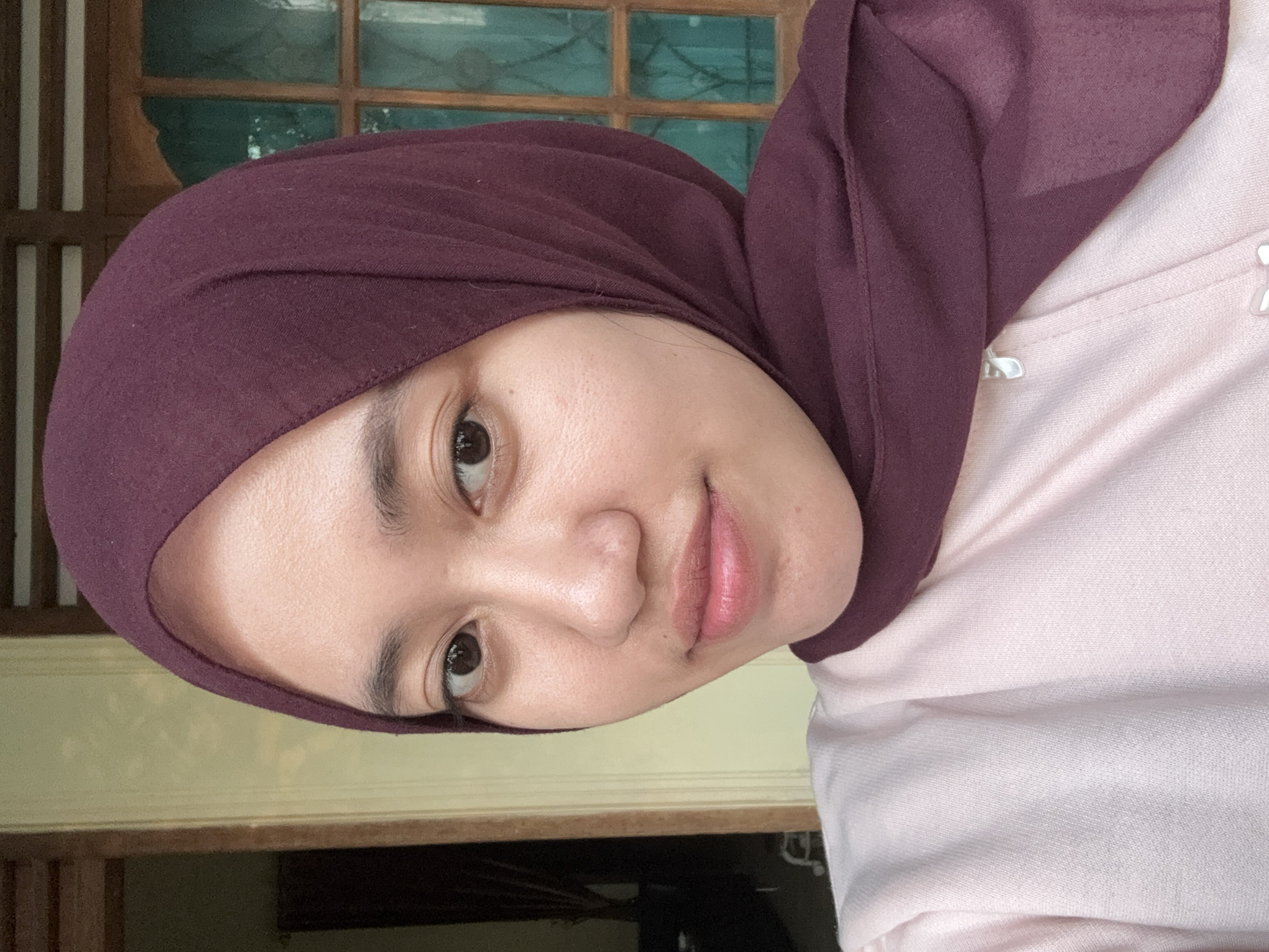 A woman is centered in the frame, angled slightly towards the right. She is wearing a dark burgundy hijab that covers her head and frames her face. She has light skin, dark eyes, and neutral-toned lips. She is also wearing a light pink shirt. The background includes a window with a wooden frame and glass panes, suggesting an indoor setting. The lighting appears soft and natural. Given the prompt's reference to Gadingrejo, Indonesia, one could infer the photo was taken in this location, though this cannot be confirmed solely from the image.