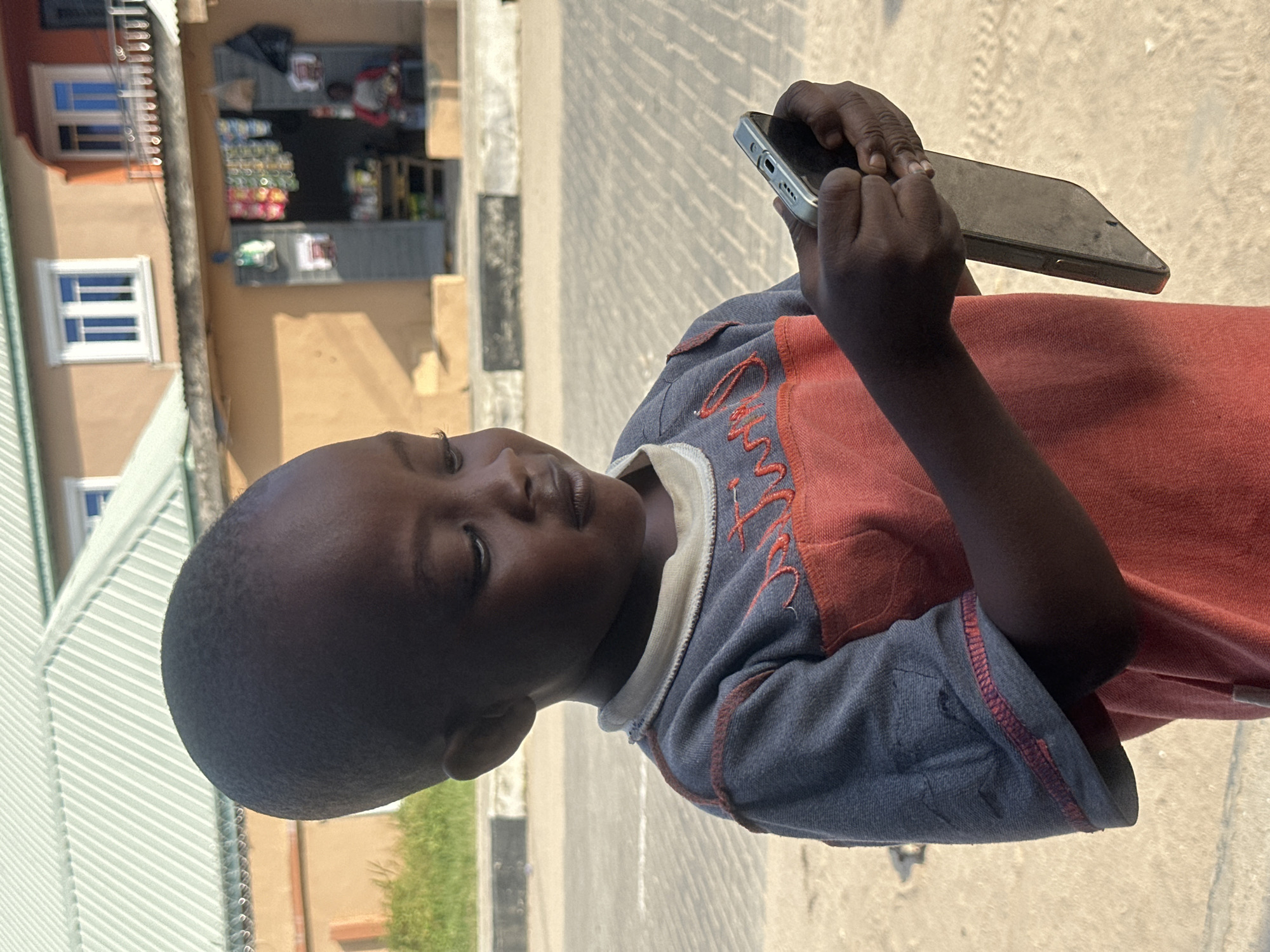 In Badagry, Nigeria, a young Black child, with short, dark hair and brown eyes, gazes intently at a mobile phone held in their hands. The child is wearing a grey and orange long-sleeved shirt with visible stitching on the seams. They stand in an outdoor environment, with a building featuring a green corrugated metal roof and orange walls in the background. This building appears to be a shop, with goods displayed in the open doorway. The ground is paved with concrete or asphalt, and a curb is visible. The lighting suggests it is daytime, with sunlight casting shadows. The child's posture and focus on the phone indicate an engagement with its content.