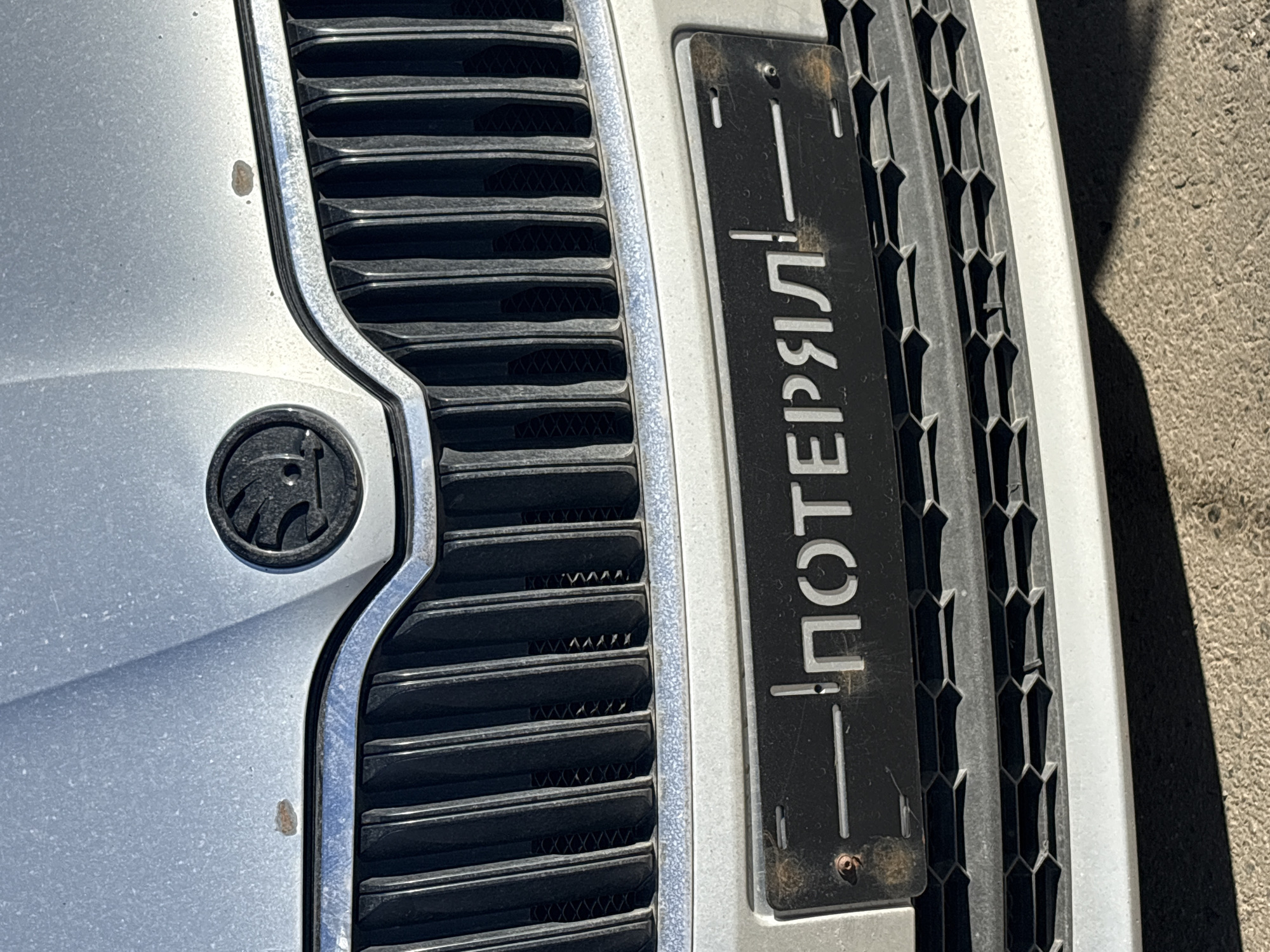 The image displays a close-up view of the front section of a car, featuring its grille, part of the bumper, and a distinctive emblem. The car is silver or light gray and appears somewhat dusty. A small rust spot is visible on the bumper, just above the grille on the left side.
A prominent feature is a black, vertically oriented rectangular sign attached to the grille on the right side. The sign displays Cyrillic text in a white or light gray stencil-like font. The text reads "ПОТЕРЯЛ!", which translates to "LOST!" or "I LOST IT!". This suggests a humorous or ironic statement, possibly indicating that the car's actual license plate is missing or that the owner is playing on the idea of losing something. The sign shows some wear, particularly around the screw holes, with hints of rust.
The car's grille has two distinct patterns: horizontal slats on the left side, and a honeycomb-like mesh pattern on the right, next to the sign. A circular car emblem, dark in color with a stylized bird or arrow design (resembling the Škoda logo), is visible on the left side of the image, positioned on the bumper near the grille.
The scene is brightly lit by direct sunlight, casting strong shadows, particularly on the ground visible to the far right. This indicates it is daytime, likely under clear skies. The ground appears to be asphalt or concrete. The Cyrillic text and implied humor are consistent with the provided location context of Izhevsk, Russia. No people or specific activities are depicted; the car is stationary.