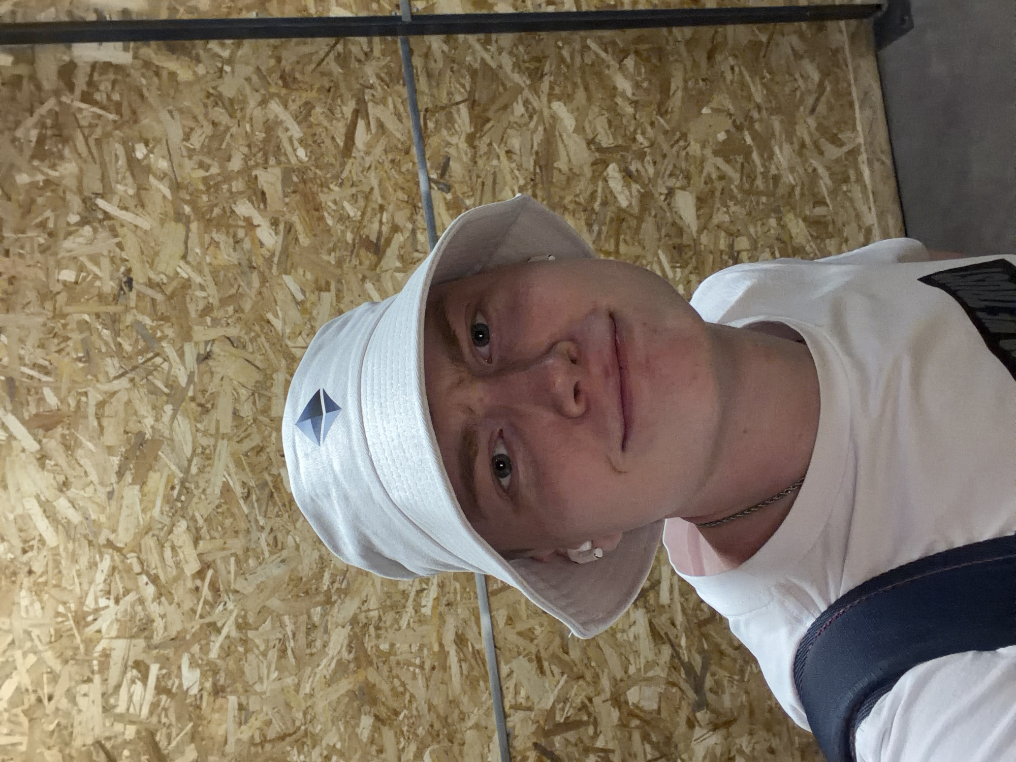 A person is photographed from the chest up, facing the camera. They are wearing a white bucket hat with a dark geometric symbol on the front, a white t-shirt, and earbuds. The background is a wall made of particle board, with a dark vertical pole to the right. The lighting is even and appears to be artificial. The location is not specified as Kyiv, Ukraine, nor is any specific city mentioned.