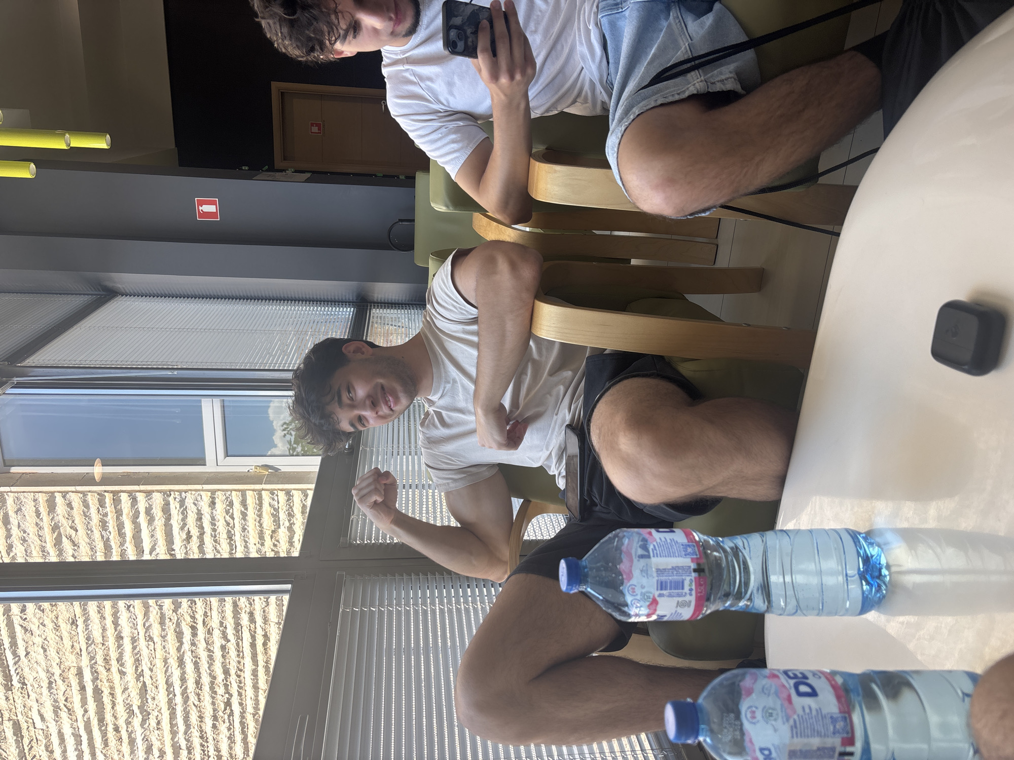 This image, rotated 90 degrees clockwise, captures an indoor scene in what appears to be a cafe or lounge, likely in Blagoevgrad, Bulgaria. Two young men are present, both with dark, curly hair and hairy legs.
In the foreground, one man, wearing a light-colored t-shirt and dark shorts, is seated, playfully flexing his biceps towards the camera with a grin. He is seated on a wooden chair with a green cushion.
Behind him, another man, dressed in a white t-shirt and light blue denim shorts, is seated in a similar chair, looking at his smartphone.
They are positioned at a light-colored table where two plastic water bottles are visible. One bottle's label clearly shows "LAVIA," and the other has "DEVIN" printed on it, both popular brands in Bulgaria. A small, black square device rests on the table near the second man.
Large windows dominate the background, fitted with horizontal blinds, allowing bright daylight to enter. Through the windows, an exterior building wall constructed from light-colored, textured stone or brickwork is visible. The strong sunlight casts noticeable shadows from the water bottles on the table, indicating a sunny day.
The interior setting includes overhead yellow tubular light fixtures and a dark wall or beam where a red fire extinguisher sign, featuring a white pictogram, is prominently displayed. The overall mood appears relaxed and casual.