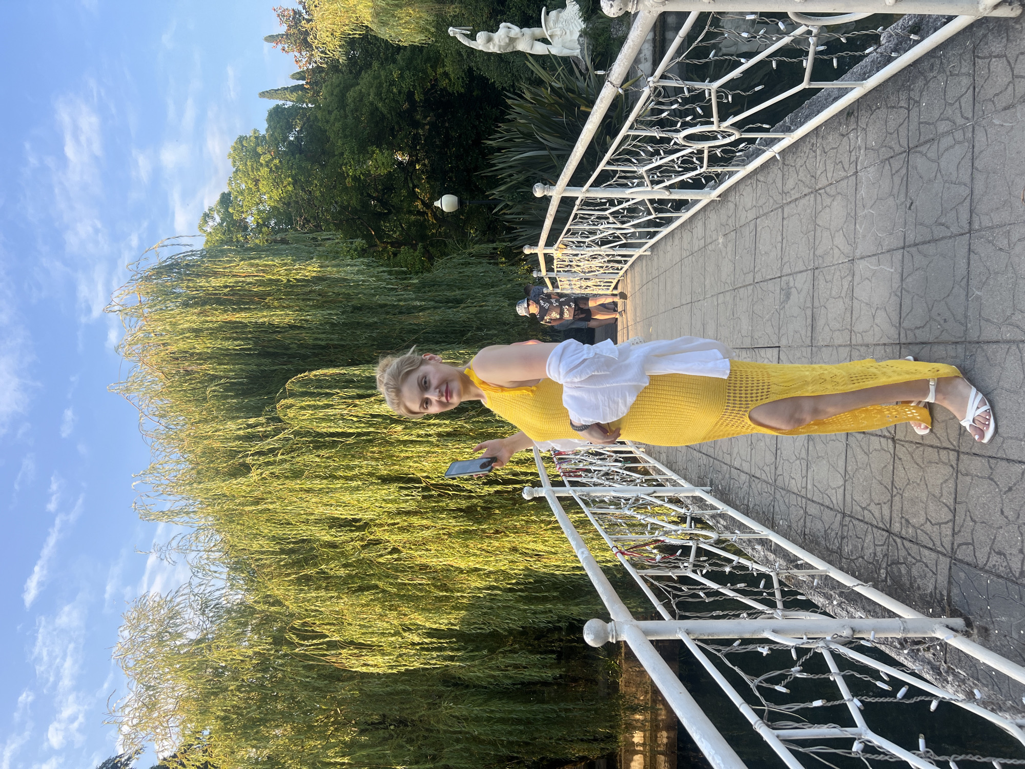 The image captures a woman standing on a white, ornate bridge in New Athos, Abkhazia. She is wearing a bright yellow, floor-length dress with a thigh-high slit and white sandals. A white shirt is draped over her shoulders. The woman is holding a smartphone in her right hand, appearing to take a photo or record a video.
The bridge spans a body of dark water, visible on either side of the walkway. The bridge's railing is decorated with what appear to be small, white string lights. Behind the woman, on the left side of the bridge, a large weeping willow tree with long, flowing green branches dominates the background, its leaves illuminated by the sun. Further in the background, trees and foliage form a dense green backdrop. To the right of the bridge, a white classical-style statue of a muscular male figure holding a spear or trident is visible, along with more lush greenery. Another person, wearing a hat and dark clothing, is walking on the bridge in the mid-ground. The sky above is a clear blue with scattered white clouds. The overall scene suggests a park or garden setting bathed in natural sunlight.